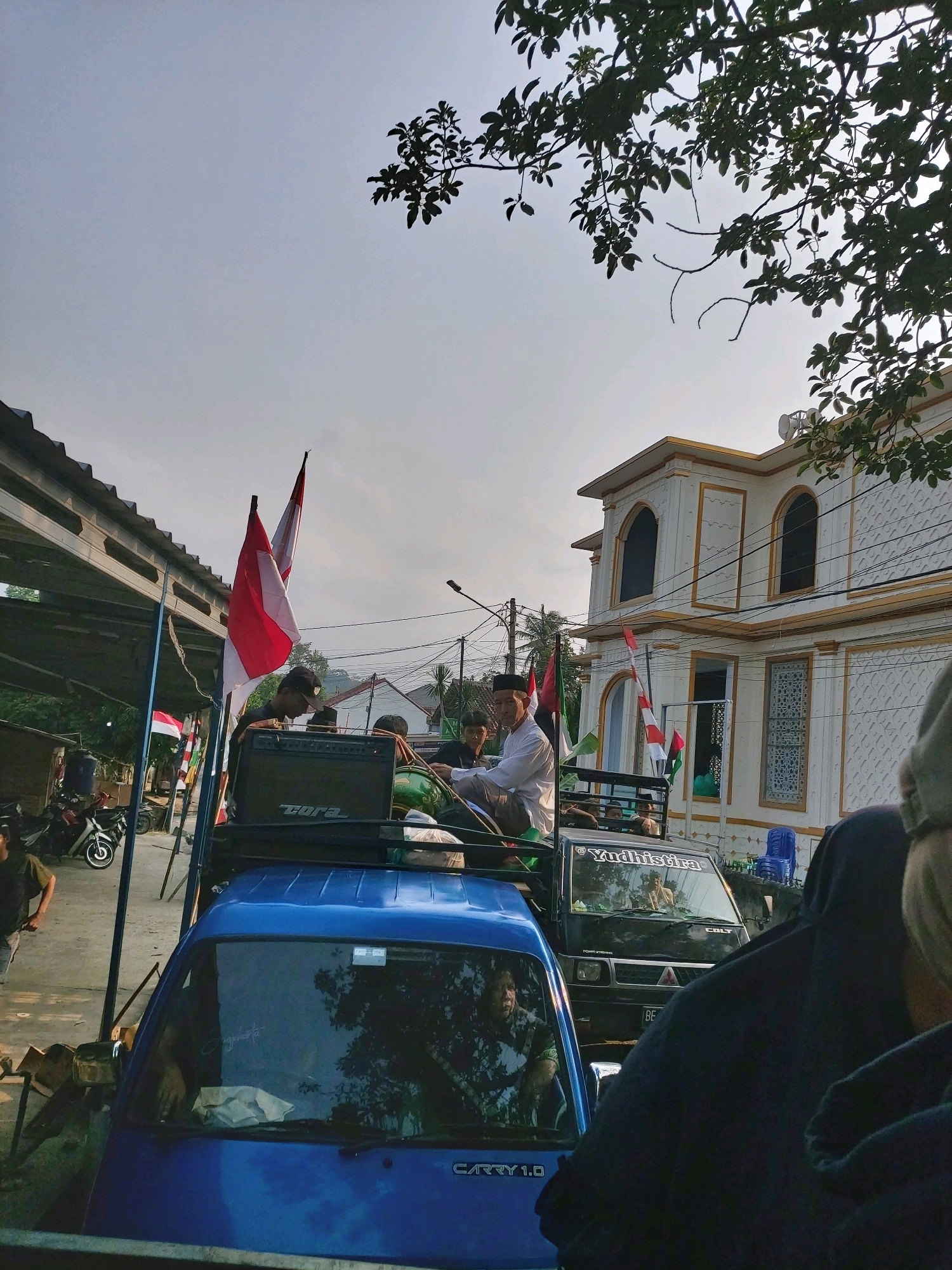 This image captures a street scene in Bandar Lampung, Indonesia, possibly during a procession or celebration, indicated by the presence of Indonesian flags. In the foreground, a bright blue pickup truck is partially visible, with its windshield reflecting trees and the sky. Behind it, a black pickup truck is parked, with several people inside and on its bed.
Dominating the view of the black truck's bed are two Indonesian flags, prominently displayed, along with a large speaker system labeled "TORA". Several individuals are visible, some appearing to be actively participating in the event. One man, wearing a cap and a dark shirt, is looking towards the speaker.
In the background, a white building with ornate gold detailing and arched windows stands out. It appears to be a place of worship or a significant structure. Trees with dark green foliage frame the top and right side of the image, suggesting an outdoor setting with some vegetation. The sky is a light gray, indicating either early morning, late afternoon, or an overcast day. The overall impression is one of a community gathering, marked by patriotic symbols and a festive atmosphere.