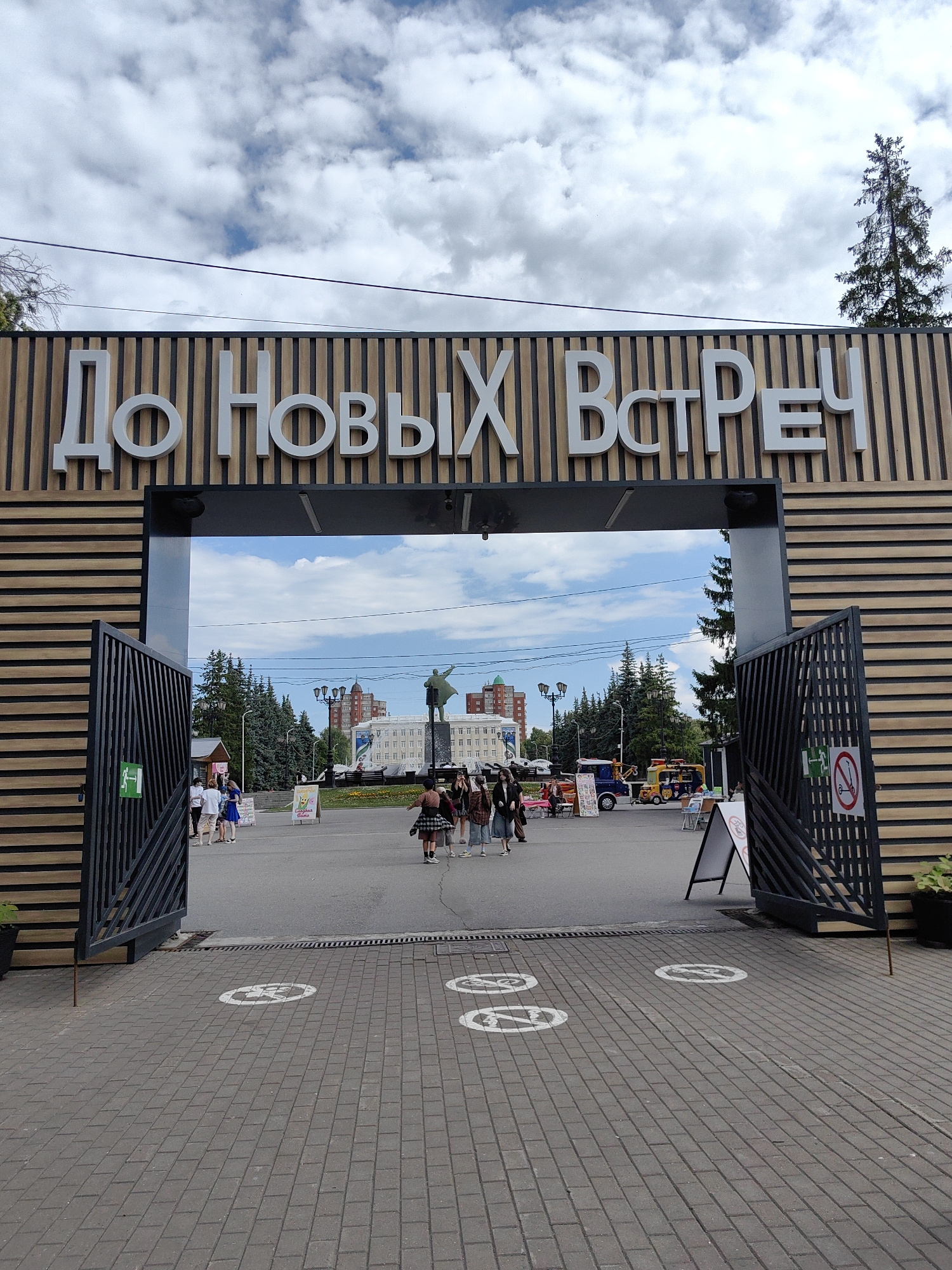 The image captures the entrance to a public space, likely a park or recreational area, in Ufa, Russia. Above the entrance, large white Cyrillic letters spell out "До Новых Встреч," which translates to "Until Next Meetings." The entrance itself is framed by a modern wooden structure with horizontal slats. To the left and right of the entrance are open black metal gates.
The scene inside the entrance reveals a paved area with several people milling about. In the background, a statue of a man with his arm raised can be seen, standing on a pedestal. Behind the statue, there are several multi-story buildings, one of which is red brick. Trees, predominantly coniferous, dot the landscape.
The ground inside the entrance is paved with grey interlocking bricks. Several circular white stencils are visible on the pavement, some of which appear to depict icons related to social distancing or designated pathways. A few small stands or carts are set up within the area, suggesting ongoing activities or vendors. The sky above is a mix of blue and white clouds, indicating daytime. The overall impression is that of an organized, well-maintained public space.
