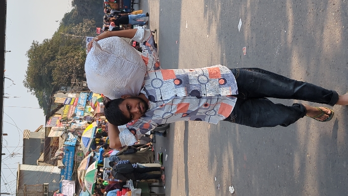 The image displays a young adult male carrying a large, light-colored sack over his right shoulder, supporting it with both hands behind his head. He is positioned centrally, looking directly at the camera, wearing a patterned long-sleeved shirt (light grey/white with orange and dark blue geometric shapes), dark blue jeans rolled at the ankles, and olive green flip-flops.
The scene is an open-air street market in Dhaka, Bangladesh. The foreground is paved ground with scattered debris. The background reveals a dense crowd of people navigating through numerous market stalls. These stalls are covered by colorful open umbrellas in shades of green, yellow, blue, and white. Behind the market, multi-story urban buildings with visible utility lines and signs bearing Bengali script are present. Large trees with dense green foliage are visible in the upper right background. The area is brightly lit by direct sunlight, indicating daytime.