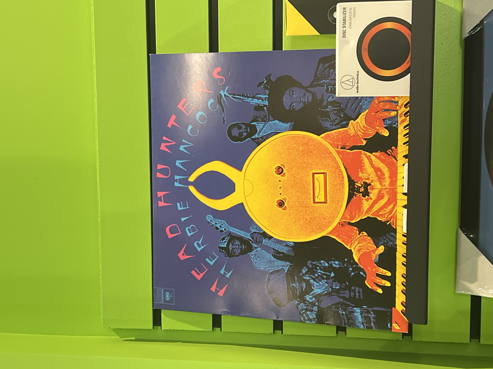 An album cover for "Herbie Hancock" (specifically "Headhunters" as indicated by "HERBIE HANCOCK" and the partially visible "HUNTERS" text) is displayed vertically against a vibrant lime green wall, likely in a record store or home setting. The album art features an illustrative style with a dark blue background. Four musicians are depicted: a central figure, presumably Herbie Hancock, with orange hands playing a keyboard; another figure to the upper left playing a saxophone; a third person holding a guitar; and a fourth, barely visible drummer in the lower right. A prominent, stylized yellow-orange object resembling a lock or a face with abstract features dominates the center of the cover. The Columbia record label logo is visible on the bottom left of the album art. Above the album, partially visible, is the packaging for an "Audio-Technica DISC STABILIZER," which also includes the French translation "STABILISATEUR DE DISQUES." The overall aesthetic of the album cover suggests a funk or jazz genre from its vibrant colors and illustrative style.
