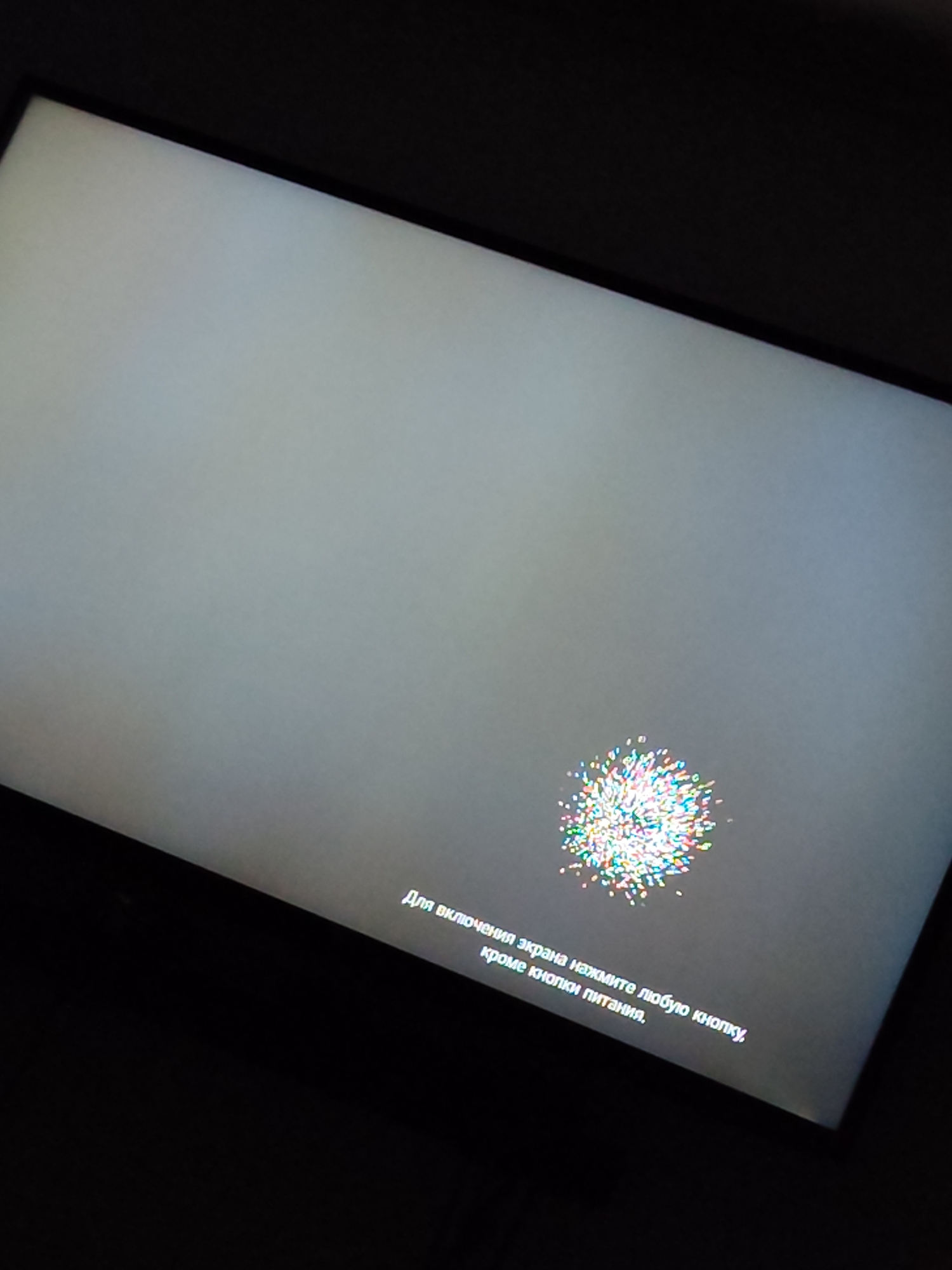 The image shows a screen displaying a message in Russian, which translates to "To turn on the screen, press any button except the power button." There is a colorful, abstract graphic near the bottom right of the screen, resembling a small firework or a cluster of confetti. The screen itself is a light gray and is angled in the shot, with the black frame of the device visible around it. The background is dark, suggesting the image was taken in a dimly lit environment. The scene is a close-up of an electronic display, with no identifiable elements of the city of Aruil de Baixo, Portugal.