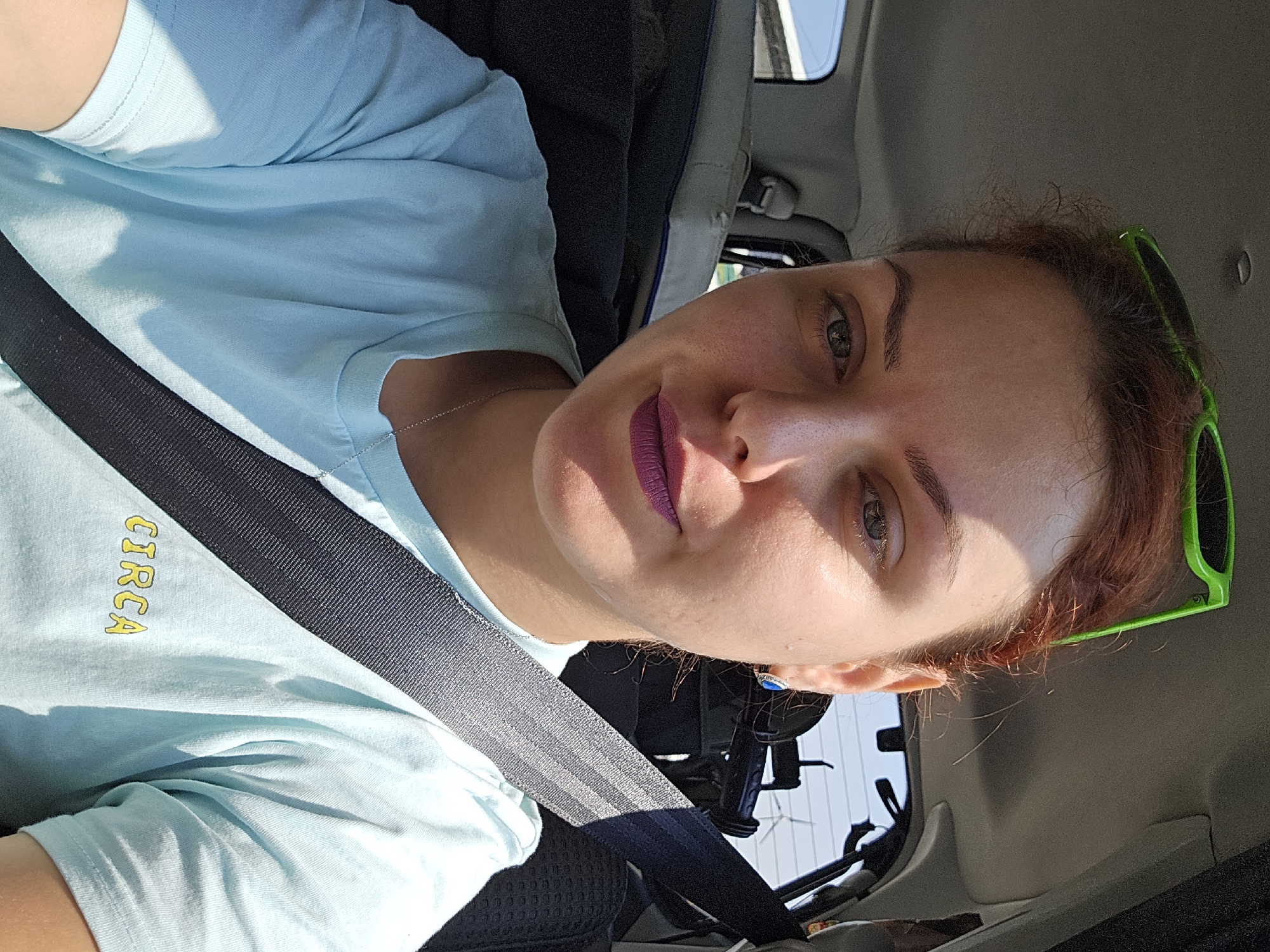 A fair-skinned woman with auburn hair, wearing bright green sunglasses on her head and a light blue t-shirt with the word "CIRCA" printed on it in yellow, is seen from the chest up, looking directly at the camera. She is wearing purple lipstick and a silver necklace. A black seatbelt is diagonally across her chest. The background shows the interior of a car, with the passenger seat and window visible. The lighting suggests it is daytime, with sunlight streaming in. The location is described as Unknown, Unknown.
