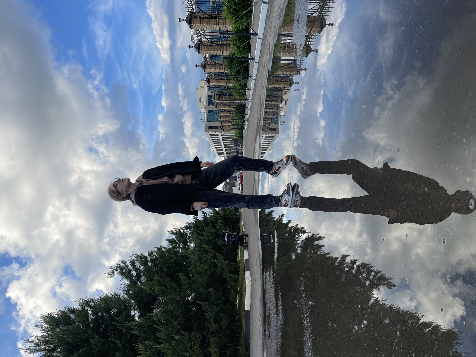 A photograph taken in Izhevsk, Russia, presents a reflection in a puddle. The reflection shows the upper portion of a building with a wrought-iron fence, a partly cloudy sky, and a person. The person is lying on the ground along the edge of the puddle, with their reflection appearing in the water. The person is wearing jeans, athletic shoes, a light-colored shirt, and a dark jacket. Trees and a road edge with black and white markers are in the foreground.
