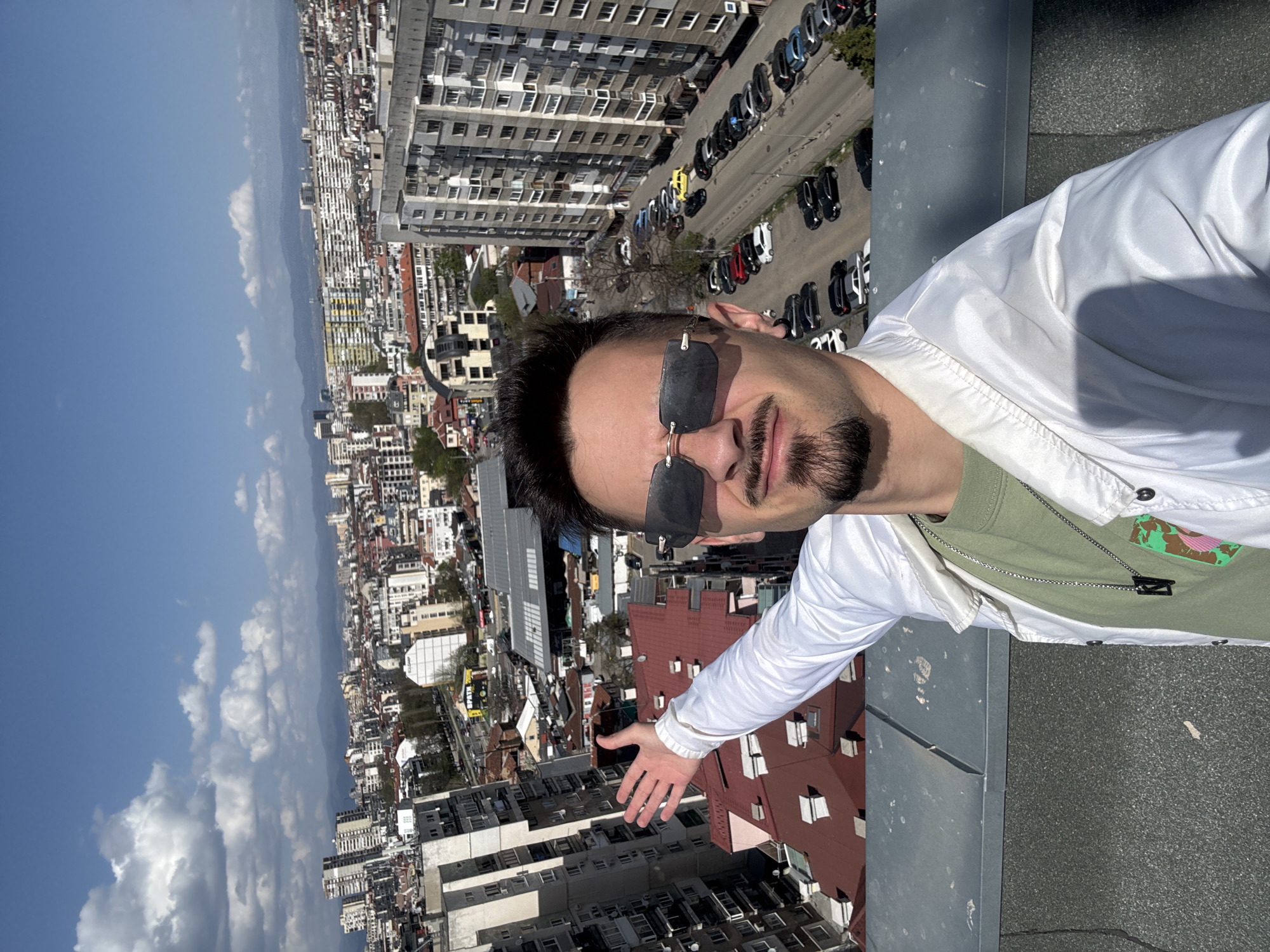A man is captured in a selfie taken from a high vantage point, likely a rooftop, overlooking the cityscape of Sofia, Bulgaria, on a clear, sunny day. The man, positioned in the foreground and facing the camera, has dark hair, a beard, and is wearing dark sunglasses. He is dressed in a white zippered jacket over a light green t-shirt, on which a patch with a stylized map-like design featuring a pink element is visible. A silver chain necklace with a black rectangular pendant hangs around his neck. His left arm is extended towards the urban panorama, with his palm open as if presenting the view.
The vast city stretches behind him under a bright blue sky dotted with scattered white clouds. The urban landscape comprises a dense array of buildings, including multi-story residential blocks, some with distinctive red-tiled roofs, and taller, more modern grey apartment structures. A street lined with numerous parked cars is visible to the man's right. In the distance, the horizon reveals a mountainous or hilly terrain. The bright sunlight casts distinct shadows, indicating midday. The foreground includes the textured surface of the rooftop and a low concrete wall.