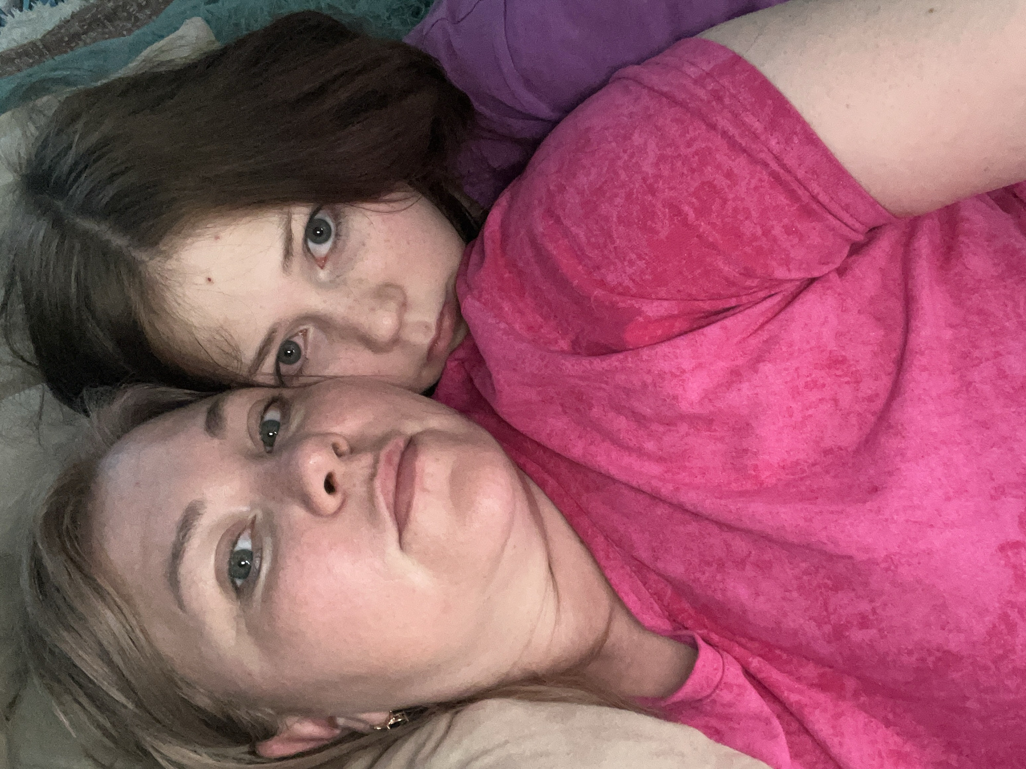 Two people, likely a parent and child, are lying down close together. The person on the bottom has blonde hair and is wearing a light-colored shirt. The person on top has brown hair and freckles, and is wearing a pink shirt over what appears to be a purple shirt. The image is a close-up selfie taken from above. There is no verifiable information to support the claim of this image being taken in the city of Vesyoloye, Russia.