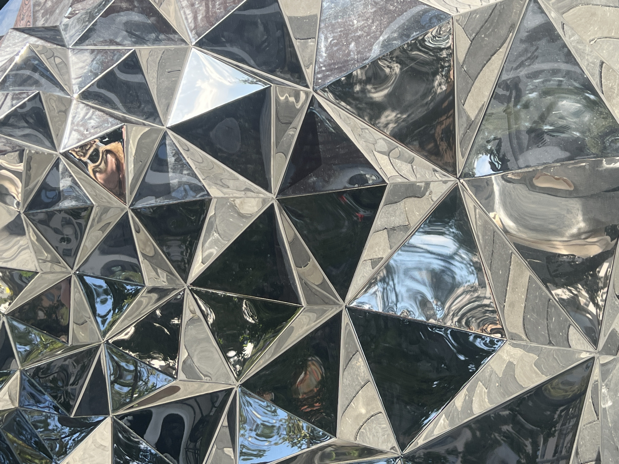 The image shows a close-up of a faceted, reflective surface, likely an architectural element or sculpture. The surface is composed of numerous triangular sections, some of which are highly polished and mirror-like, reflecting the surrounding environment. These reflections show distorted images of trees, sky, and possibly people. Other sections of the surface appear to be matte or have a textured finish, creating a contrast with the reflective parts. The overall impression is one of modern design and geometric complexity. The lighting suggests it is daytime, with sunlight casting reflections and highlights. The location context provided is Yekaterinburg, Russia, but there are no specific visual cues within the image to confirm this. No text is visible in the image.
