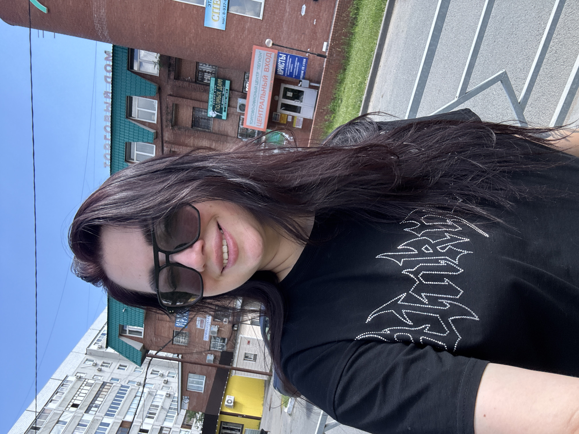 A young woman with long, dark hair and sunglasses smiles at the camera, taking a selfie outdoors. She is wearing a black t-shirt with a white, fiery graphic design. In the background, a multi-story brick building with a green roof is visible, along with a tall brick tower. The building has several windows and signs, one of which reads "ТОРГОВЫЙ ДОМ" (Trading House). Another sign indicates "ЦЕНТРАЛЬНЫЙ ВХОД" (Central Entrance). A large apartment building with many windows is also present in the background. The scene appears to be in an urban setting under a clear blue sky, with a paved area and a metal fence in the foreground.