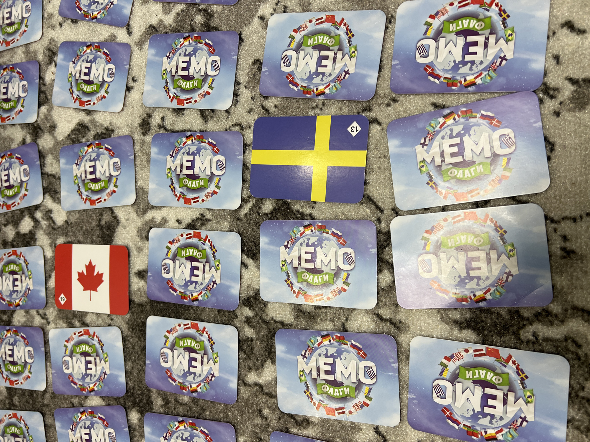 The image shows a collection of cards scattered on a gray carpet with a slightly blurred pattern. Most of the cards feature a circular design with various national flags surrounding the word "MEMO" (ФЛАГИ). Two cards are different: one displays the Canadian flag with a maple leaf, marked with the number "19", and the other shows the Swedish flag with a yellow cross on a blue background, marked with the number "13". The scene is indoors, and the objects appear to be part of a memory matching game. There are no people visible in the frame. The image has been taken with an angle looking slightly down on the scene. There are no clear markers indicating a specific location in Chaikovsky, Russia.