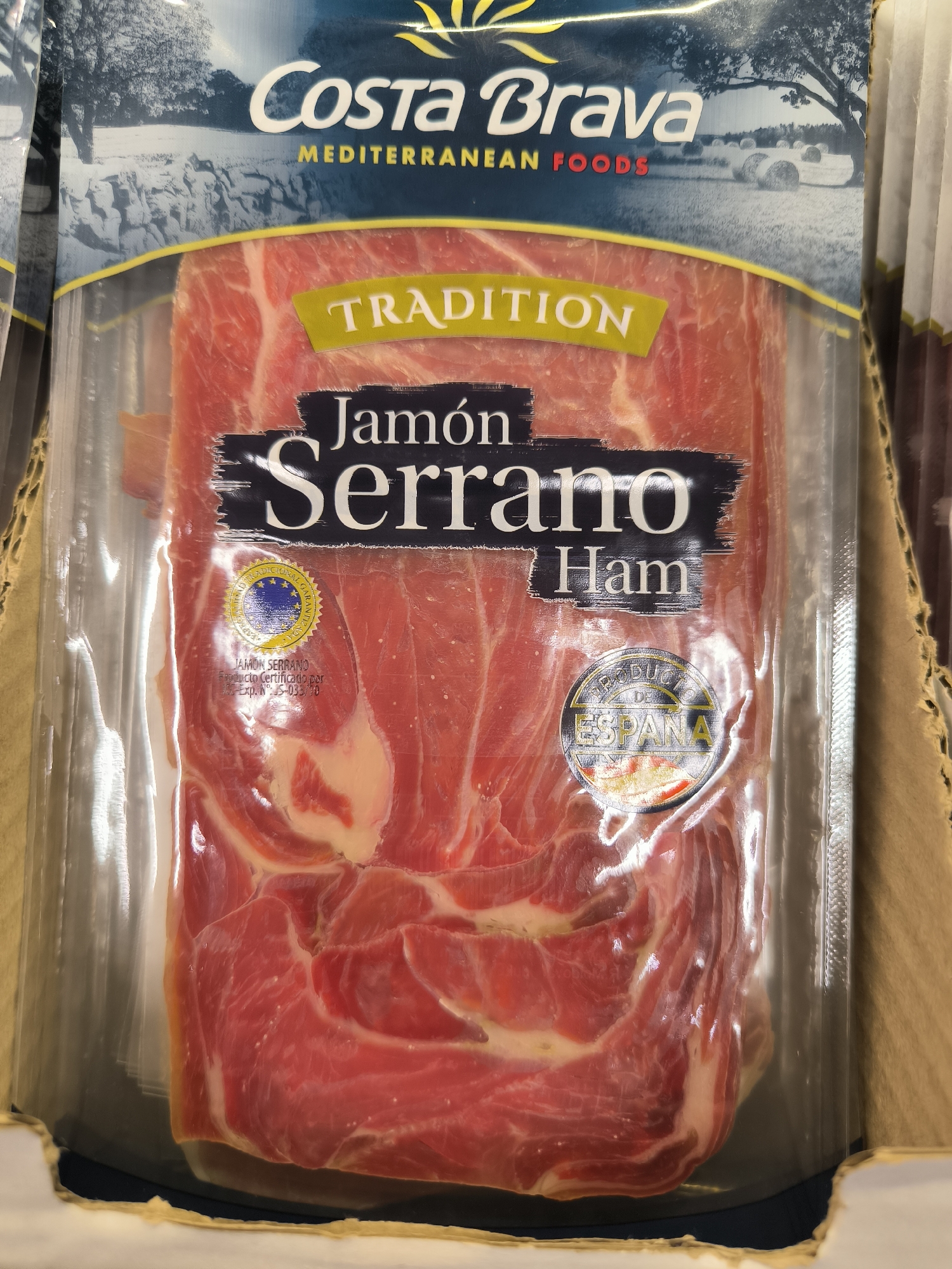 A package of Costa Brava Jamón Serrano Ham is displayed in a retail setting. The packaging is clear plastic, revealing thinly sliced cured ham. Text on the packaging includes "Costa Brava," "MEDITERRANEAN FOODS," "TRADITION," "Jamón Serrano," and "Ham." There are also circular seals indicating "PRODUCTO CERTIFICADO" and "DE ESPAÑA," and a "Exp. Nº. ES-033/70." The ham is arranged in overlapping slices within the package. The background shows shelves and other products, typical of a supermarket or specialty food store. The lighting is artificial, likely from overhead store lights. This item is not specifically located in Chișinău, Moldova.