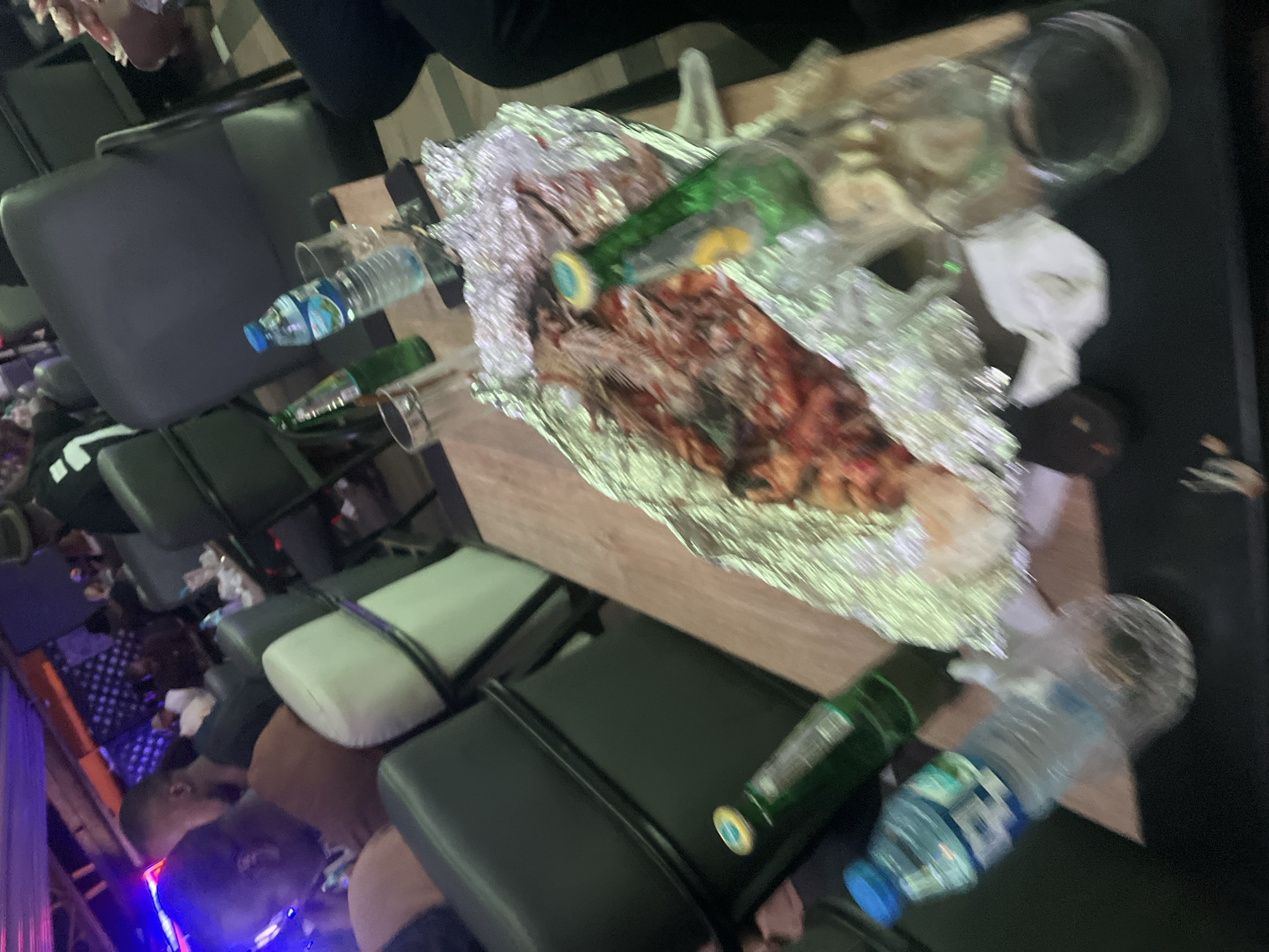 This image captures an indoor scene within a restaurant or bar in Abuja, Nigeria. The environment is dimly lit, featuring dark upholstered chairs and wooden tables. Artificial lighting in the background casts purple and orange hues. In the foreground, a light-colored wooden table displays a partially consumed meal, likely grilled fish or meat with vegetables and sauce, presented in an open aluminum foil wrap. Visible within the foil are scattered bones, indicating consumption. Alongside the food are two green glass bottles (one with a yellow/gold cap, one mostly empty), two plastic water bottles with blue caps (one nearly full, one partially consumed), and several clear drinking glasses, some empty. A black mobile phone is also present on the table. In the mid-left foreground, a person wearing a brown or orange shirt is visible from the side, seated. To the immediate right, a leg clad in dark trousers is partially visible. Further in the background, several other patrons are seated at various tables, appearing to be dining and socializing in a casual setting.