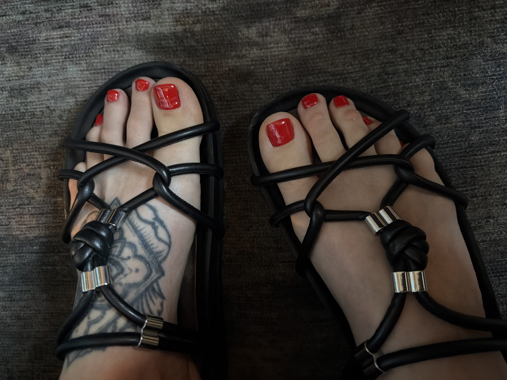 The image shows a view of a person's feet clad in black gladiator-style sandals with red painted toenails. The sandals feature a crisscrossing strap design with decorative knots and silver metallic accents. The right foot exhibits a tattoo on the instep, primarily in black ink. The background consists of a dark, textured surface, possibly a rug or carpet. The location is potentially in West Hollywood, United States of America, though there are no readily identifiable landmarks within the frame. No specific actions or interactions are visible.