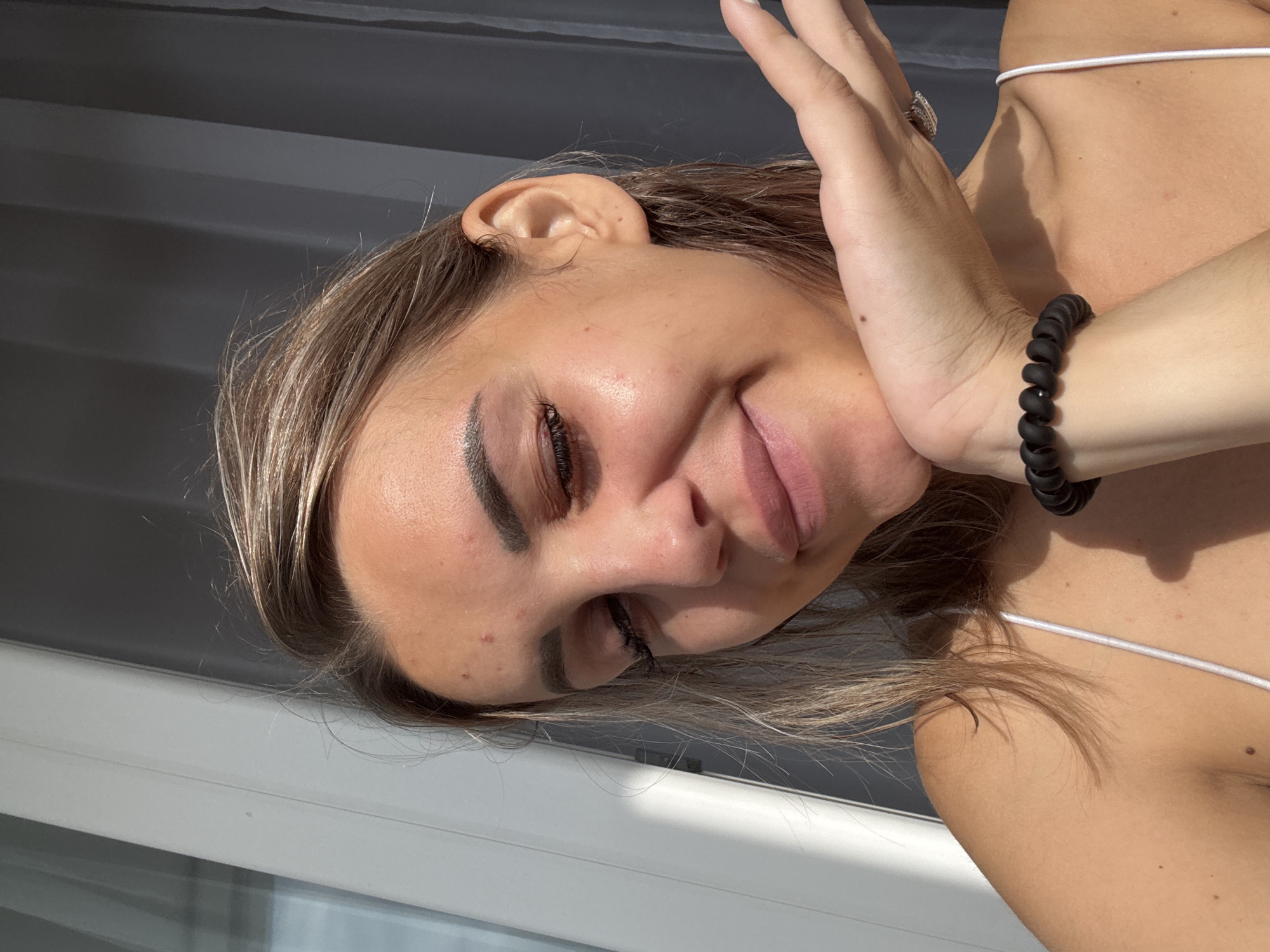 A woman is posing for a selfie in what appears to be a brightly lit indoor setting with sheer curtains in the background. She has her hand resting on her chin and is looking downwards with a slight smile. She is wearing a white tank top and a black coiled bracelet on her wrist. The lighting suggests it is daytime. No specific location in Kholmsk, Russia can be determined from the image.