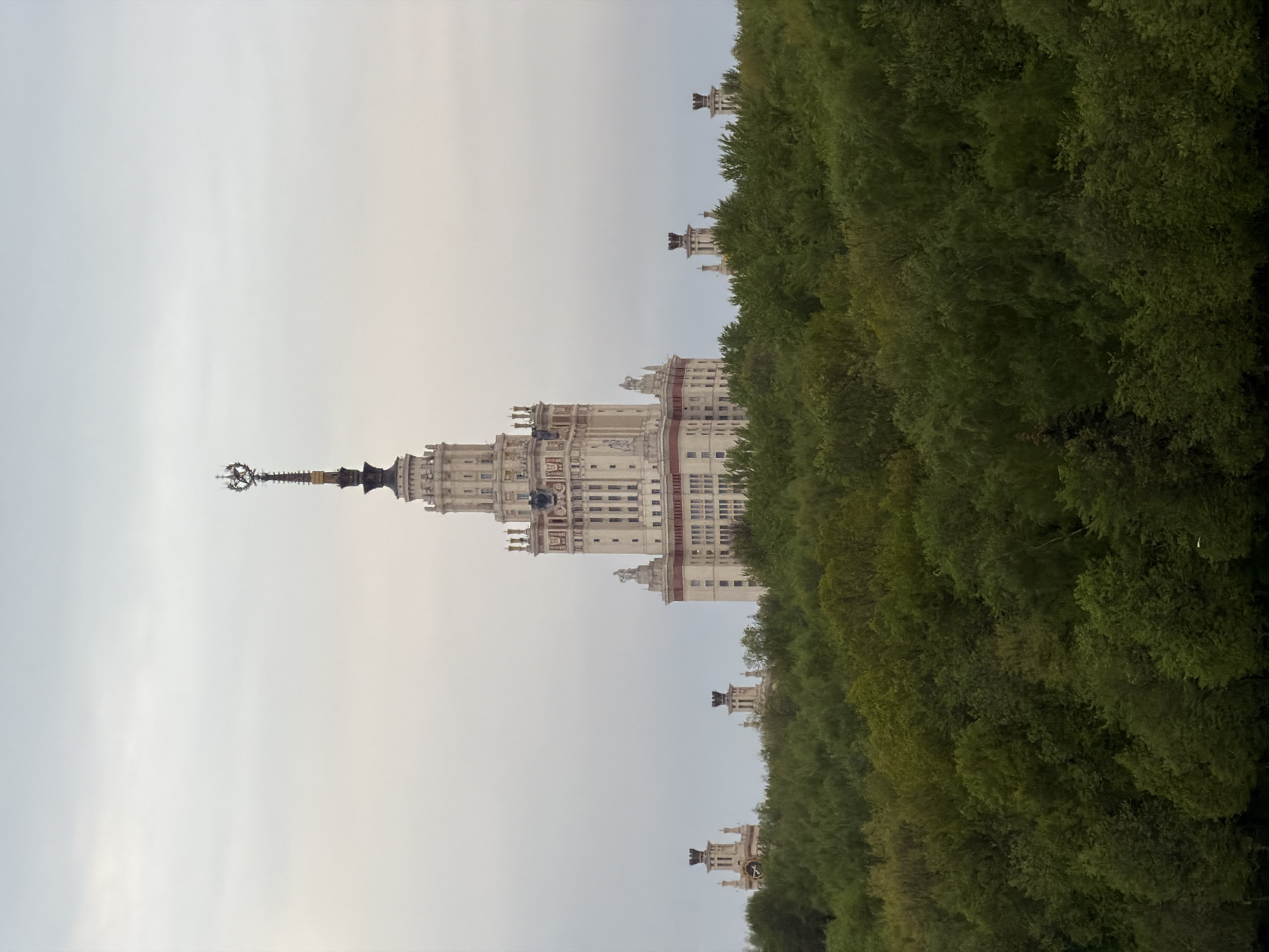 The image shows the Moscow State University building in Moscow, Russia. The building is a large Stalinist skyscraper, with a central tower and multiple wings. The architectural style is characterized by its monumental scale, classical detailing, and a spire topped with a star. The building is partially obscured by green trees on the right side. The sky is overcast and gray.