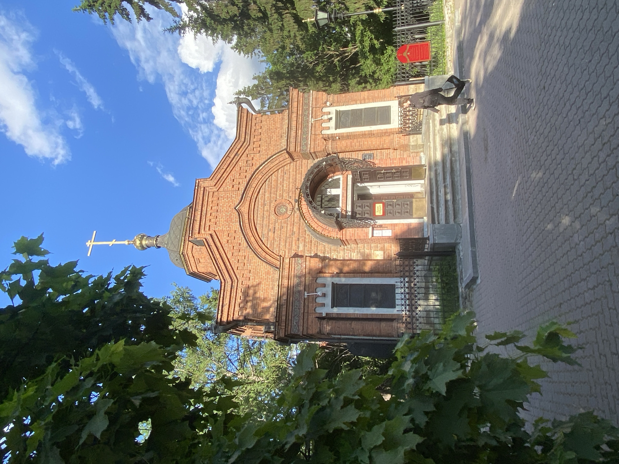 The image depicts the Church on the Blood in Yekaterinburg, Russia, viewed from a low angle through foliage. The church, made of red brick, features a golden dome topped with a cross. A person is walking along the paved pathway adjacent to the church. The sky is blue with scattered clouds. A lamppost and a red sign are visible near the church's entrance.