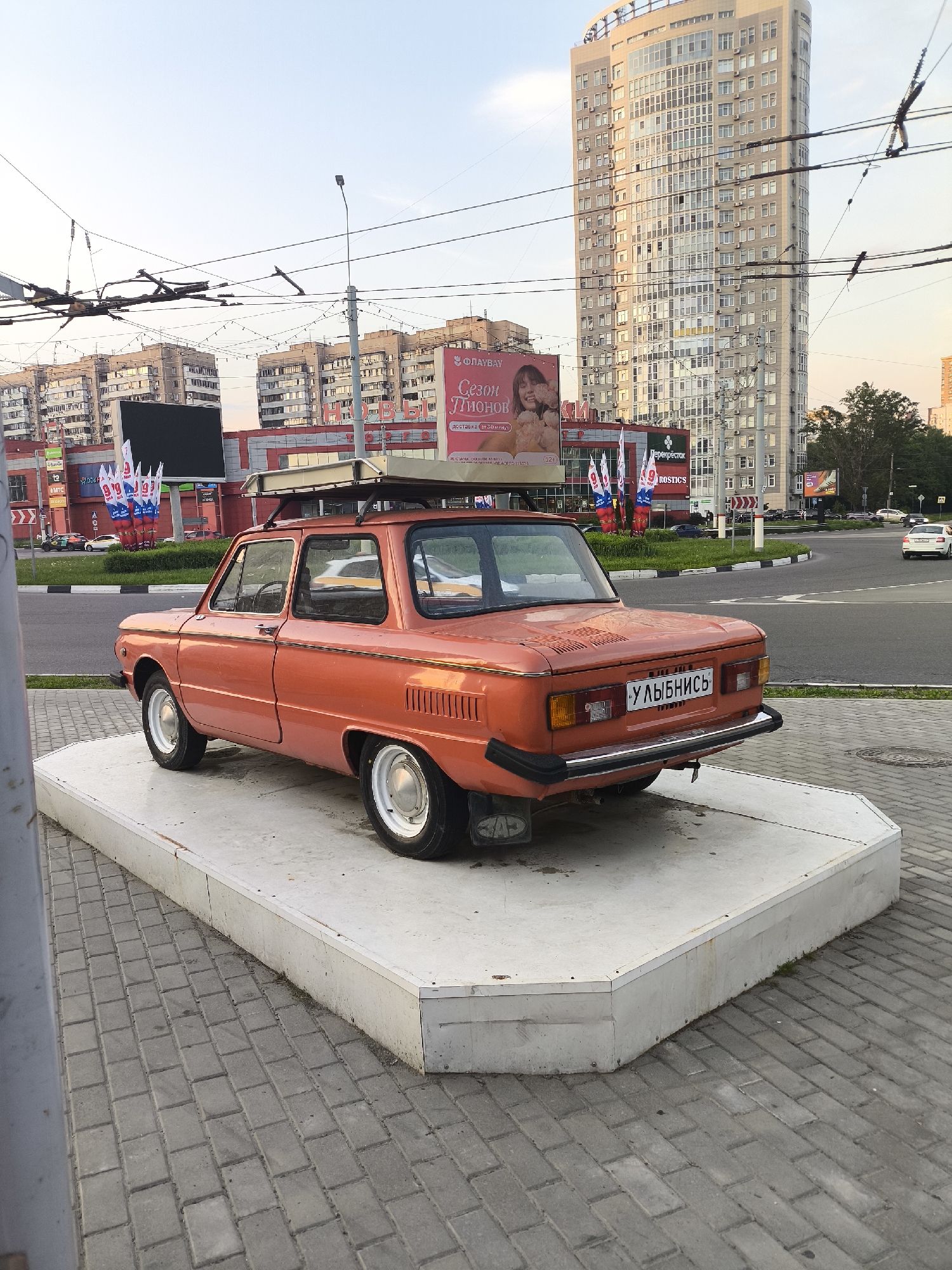The image depicts an orange ZAZ-968M car mounted on a white, two-tiered platform. The car has a luggage rack on its roof and a license plate that reads "УЛЫБНИСЬ" (smile). The setting is an urban area, with buildings, a shopping center, a tall skyscraper, and street infrastructure visible in the background. The sky is light blue, indicating it could be late afternoon or early evening.
The car and platform are situated on a paved area with gray bricks, near a road and a roundabout. There are light poles with overhead cables visible above. The buildings in the background include a shopping center with signs for "Перекрёсток", "ROSTIC'S" and a billboard advertising flowers. There is also a tall residential building in the background.
There are some cars on the road, indicating that the scene is a typical urban setting in an unknown city. There are some flags with the number "9" on them.