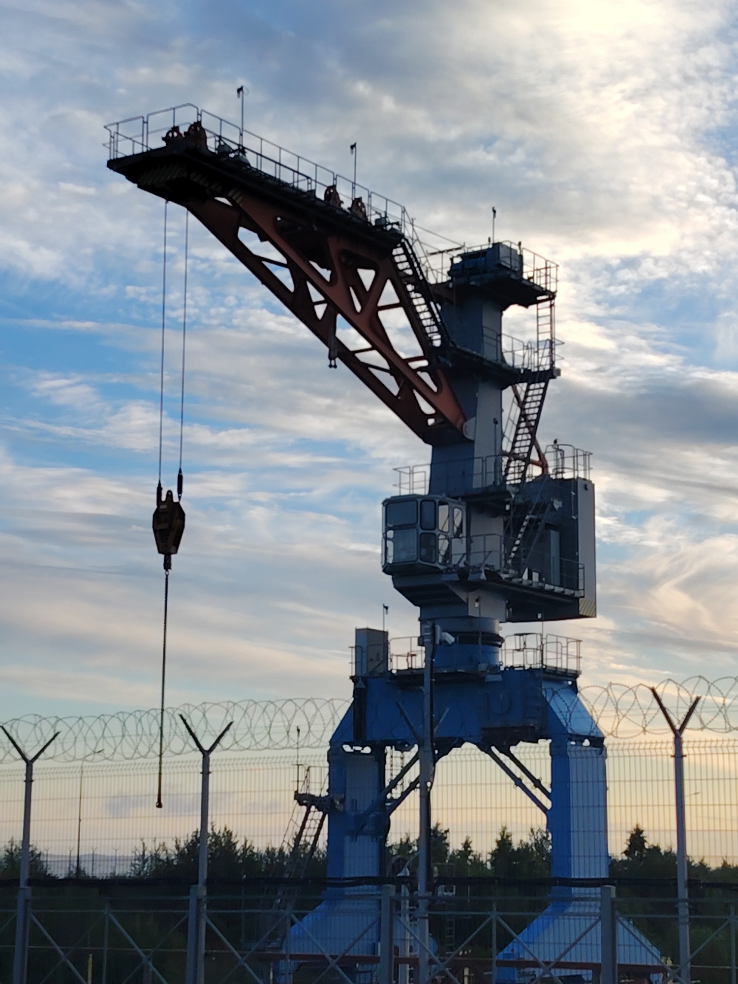A large, blue and red gantry crane stands silhouetted against a cloudy sky at the Chaikovsky Combine in Chaikovsky, Russia. The crane has a long, angled boom with a hook hanging from a cable. A metal fence with barbed wire runs across the foreground, partially obscuring the base of the crane and a line of trees behind it. The sky is a mix of blue and white clouds, with some hints of orange from what appears to be sunset. There are no people visible in the image.