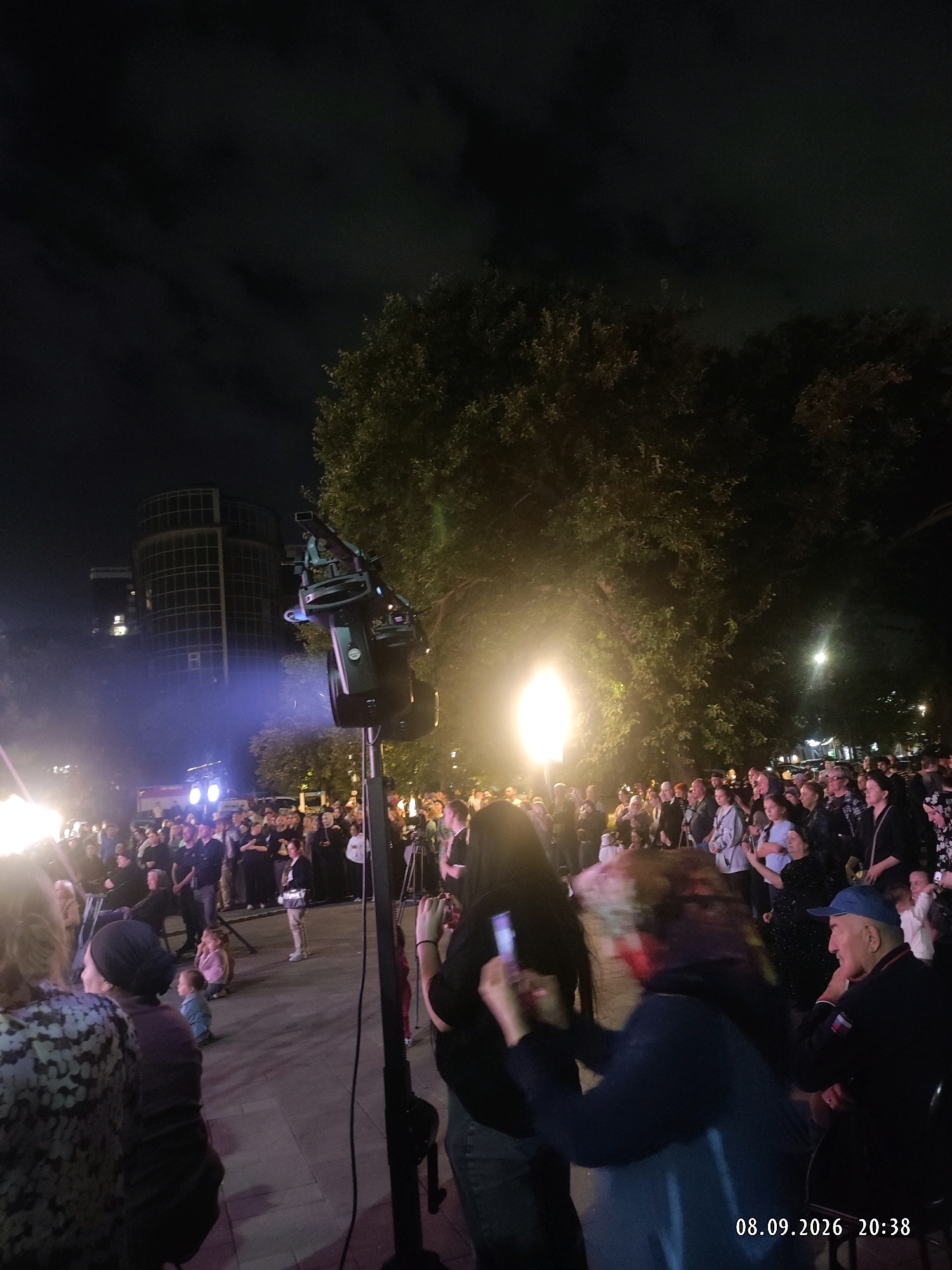 An outdoor night event is depicted with a crowd of people, some of whom are holding up phones to record. In the foreground, a large piece of equipment, possibly a camera rig or lighting equipment, is mounted on a stand. Further back, a brightly lit area suggests a stage or focal point. A building with a modern architectural design is visible in the background. The overall scene suggests a public gathering or performance. The date stamp on the image indicates it was taken on September 8, 2026, at 20:38.