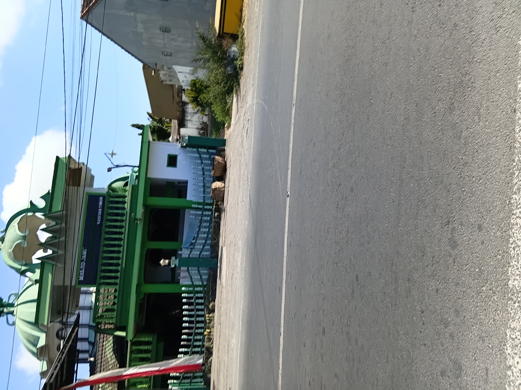 The image shows a street view in Kasiyan Timur, Indonesia. In the background, a green mosque with white trim and domes is visible. A white and red flag is displayed on the left side of the mosque. A green fence runs along the front of the mosque. A paved road with a white line marking is in the foreground. There are some baskets on the sidewalk in front of the mosque. The sky is clear blue with some clouds. The overall scene depicts a typical street in a residential or commercial area with a prominent religious building.