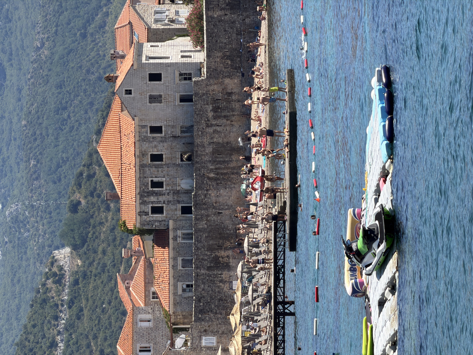 The image captures a vibrant scene at the beach in Budva, Montenegro. The foreground is dominated by the clear blue water, with a floating platform featuring a jet ski and inflatable toys. A rope barrier with buoys separates the swimming area from the deeper water. In the mid-ground, a sandy beach is packed with sunbathers relaxing on lounge chairs under umbrellas. A pier extends into the water, with several individuals standing and walking on it. Behind the beach, a rustic stone wall forms a barrier, beyond which lie several buildings with characteristic orange-tiled roofs, typical of the Budva Old Town architecture. In the background, a lush green mountain slopes upwards, providing a scenic backdrop to the coastal town. The atmosphere is one of a busy summer day, with many people enjoying the sun, sea, and recreational activities.