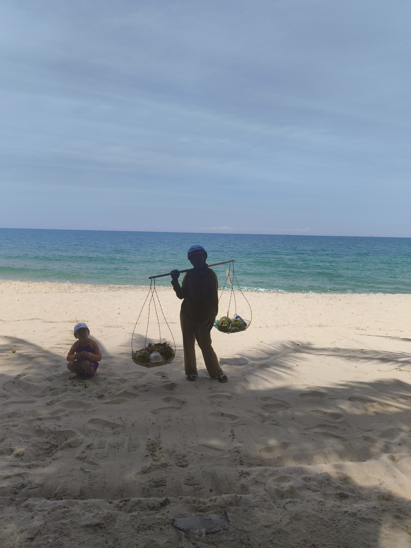 An eye-level, full outdoor shot captures a beach scene in Phu Thuy Ward, Vietnam. The foreground shows sandy beach with visible footprints and palm tree shadows. In the middle ground, a person is crouched on the left, wearing a purple and orange long-sleeved shirt, purple shorts, and a white hat. To the right, a person stands wearing a dark long-sleeved shirt, dark pants, a helmet, and gloves. They are carrying a yoke across their shoulders, from which two baskets are suspended. The left basket contains various fruits, and the right basket holds several coconuts. The background features the turquoise ocean meeting the horizon under a clear blue sky with light clouds.