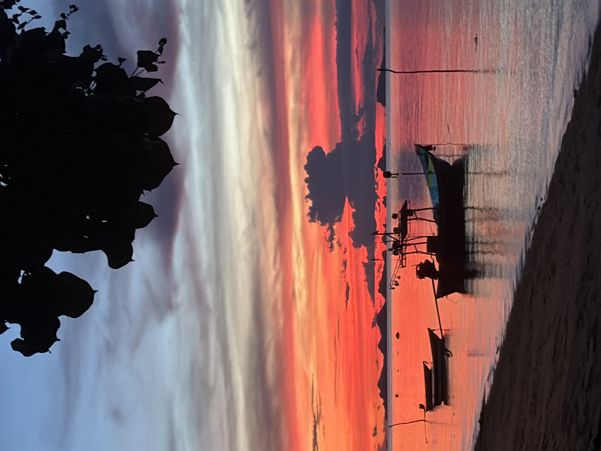 This is a vertical, eye-level, full-shot photograph taken at Baan Thong Po, Thailand, during sunset. The sky transitions from a pale blue at the top to vibrant shades of orange and red near the horizon, with streaks of gray and pink clouds scattered throughout. Silhouetted against the colorful sky are several islands in the distance.
In the foreground, a dark, sandy beach meets the calm ocean. The water reflects the fiery hues of the sunset, creating a shimmering surface. Two small, dark-colored boats are anchored in the shallow water near the shore. Further out, a larger fishing boat with a dark silhouette is also anchored. A thin, dark pole, possibly part of a dock or marker, stands vertically in the water to the right of the fishing boat.
The upper portion of the image is dominated by the dark silhouette of tree leaves and branches, which hang over the scene, framing the sunset and the water below. No people are visible in the image. The overall atmosphere is serene and tranquil.