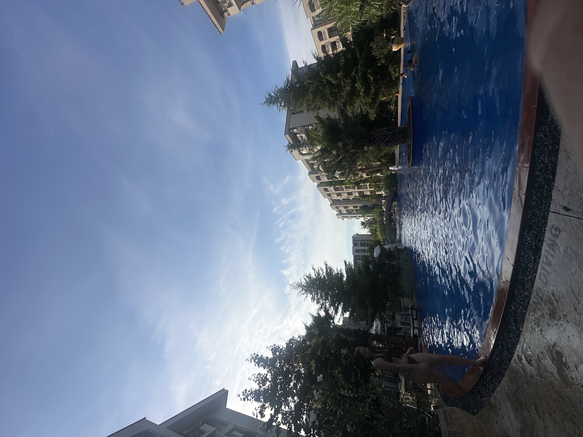 A wide-angle shot captures an outdoor swimming pool area with several buildings and lush trees surrounding it under a partly cloudy blue sky. In the foreground, to the left, a woman in a bikini stands at the edge of the pool, with her feet in the water. The pool itself is a vibrant blue, with gentle ripples on the surface. In the middle ground, several people are visible swimming and relaxing in the water. A large, circular planter with a palm tree is situated in the pool. Behind the pool, a series of multi-story buildings with balconies are visible, interspersed with tall evergreen trees and some palm trees. The architecture suggests a resort or hotel complex. The overall scene depicts a sunny day at a recreational facility, likely a resort or hotel.