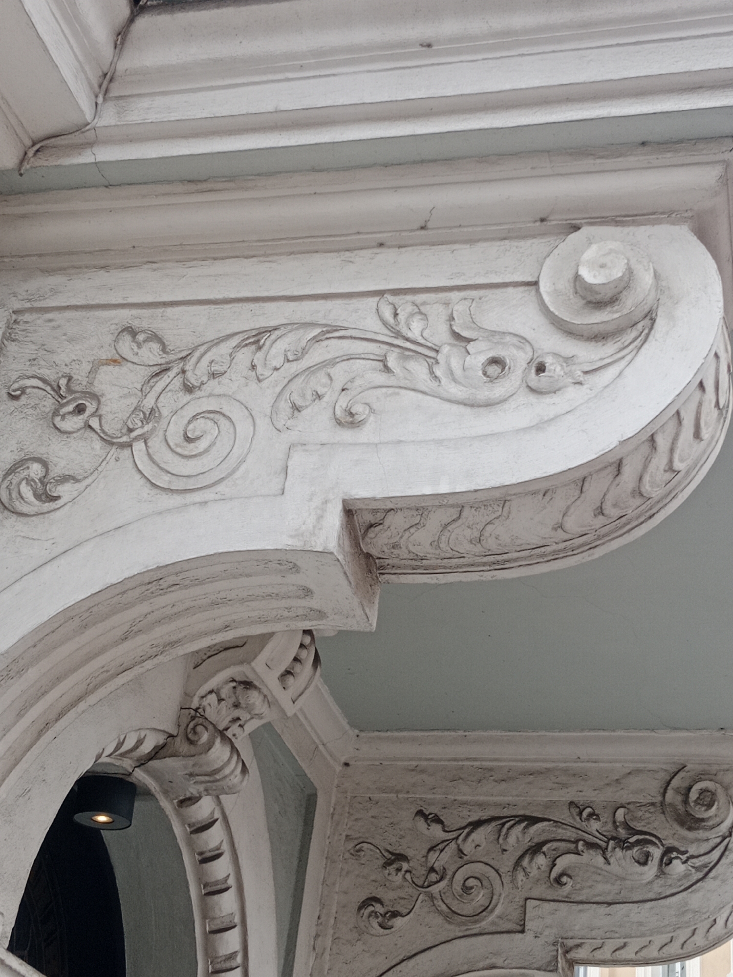 The image shows architectural details of an ornate facade, likely in Bydgoszcz, Poland. The subject is an exterior view of decorative plasterwork. The plasterwork is in a pale, off-white color and features intricate scrolling floral motifs, volutes, and acanthus leaves. There is evidence of age and some minor cracking in the plaster. The architectural element is curved and appears to be part of a larger structure, possibly an entryway or the underside of a balcony. A modern cylindrical light fixture with a warm orange glow is visible within a recess to the left of the frame. The background consists of a light blue ceiling or overhang. The style suggests historical architecture, possibly Beaux-Arts or Art Nouveau, common in European cities.