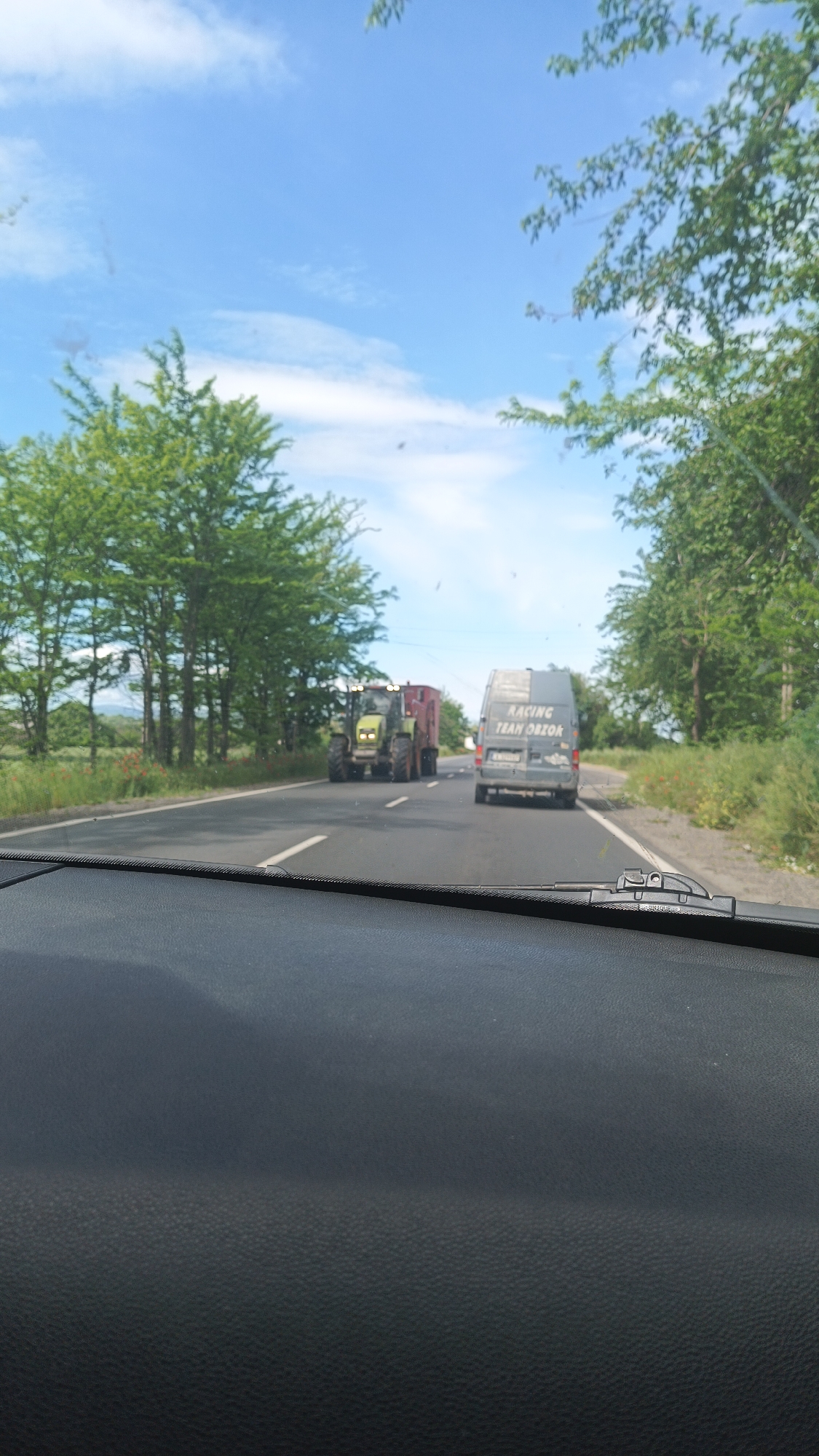 The image depicts a road scene from inside a vehicle, likely a car. The dashboard is visible in the foreground. The road is straight, marked with white lines and asphalt, leading into the distance. On the road, there's a green tractor pulling a red trailer, followed by a grey van with "RACING TEAM ORZOK" written on the back. The surrounding environment includes green trees lining both sides of the road, suggesting a rural or semi-rural setting. The sky is blue with some white clouds. Some red flowers are visible in the grass next to the trees. The city of Popovitsa, Bulgaria is assumed to be the location.