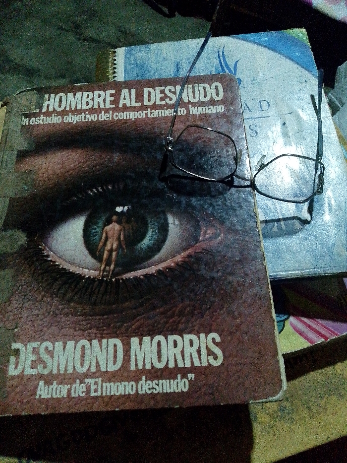 The image shows two books and a pair of eyeglasses. The top book, titled "El Hombre al Desnudo," features a close-up of an eye with a nude figure standing in the pupil. The author is listed as Desmond Morris, known for "El Mono Desnudo." The second book is partially visible, appearing older and possibly containing illustrations. The eyeglasses are modern, with a thin metal frame and rectangular lenses, casually resting on top of the books. The lighting is dim, casting shadows and obscuring some details.