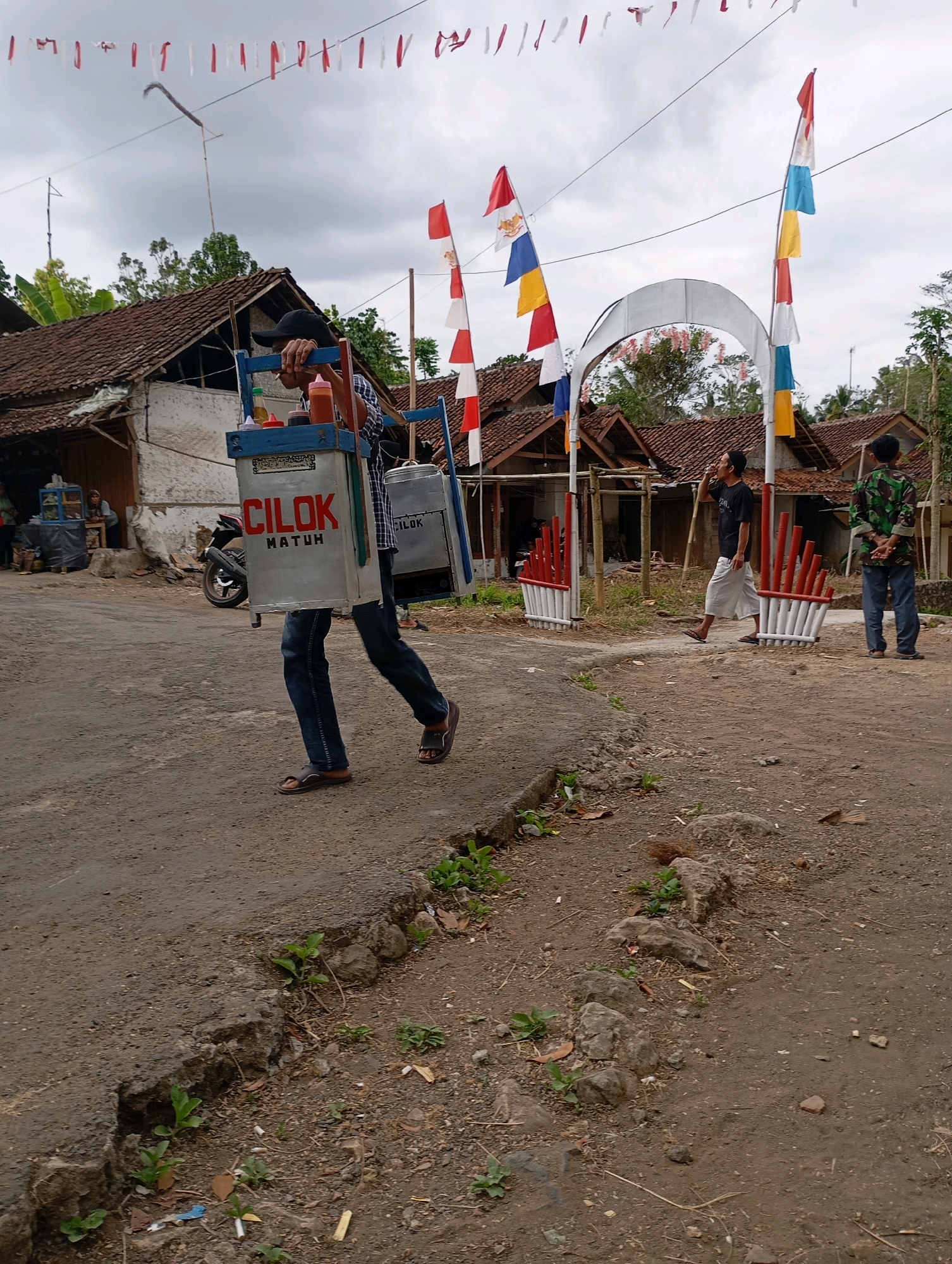 In Cigunung, Indonesia, a man carrying two large metal containers, labeled "CILOK MATUH", walks along a dusty road. The containers appear to be food carriers, likely for selling snacks. He wears a plaid shirt, dark pants, and sandals. Behind him, an arched gateway decorated with red and white striped flags stands. Two other men are visible; one in a black t-shirt and white pants stands near the gateway, and another in a camouflage shirt and blue jeans stands further in the background. The scene is set against a backdrop of traditional Indonesian houses with tiled roofs and lush green trees under an overcast sky. Several red and white flags, along with colorful triangular flags, are strung across the scene, suggesting a local celebration or event. A motorcycle is parked near one of the houses, and various small plants grow along the roadside.
