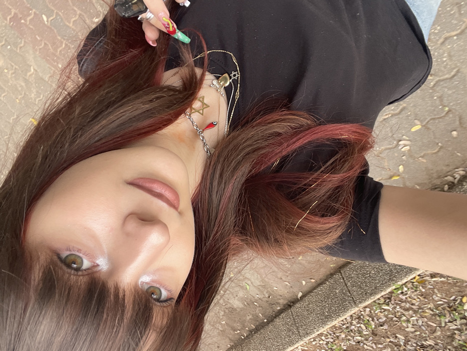 A selfie depicts an individual outdoors in Raanana, Israel. The subject, appearing female, possesses long, dark brown hair with prominent red/burgundy highlights and thin golden tinsel strands, styled with bangs covering the forehead. Her face features light brown/hazel eyes with white inner corner eyeliner and light pinkish-brown lipstick, exhibiting a subtle smile. She wears a black t-shirt. Multiple layered necklaces adorn her neck: a silver chain with a small red teardrop-shaped charm and a small silver Star of David pendant, alongside a longer gold chain with a larger gold Star of David pendant and a gold heart-shaped charm. Her partially visible right hand displays several rings and elaborately painted, long fingernails, which include green, red, and patterned designs. The immediate environment is a paved area constructed with light brown interlocking blocks, bordered by a concrete curb adjacent to a strip of ground covered in scattered dry brown and yellow leaves. The specific location within Raanana is not discernible from the visual information provided.