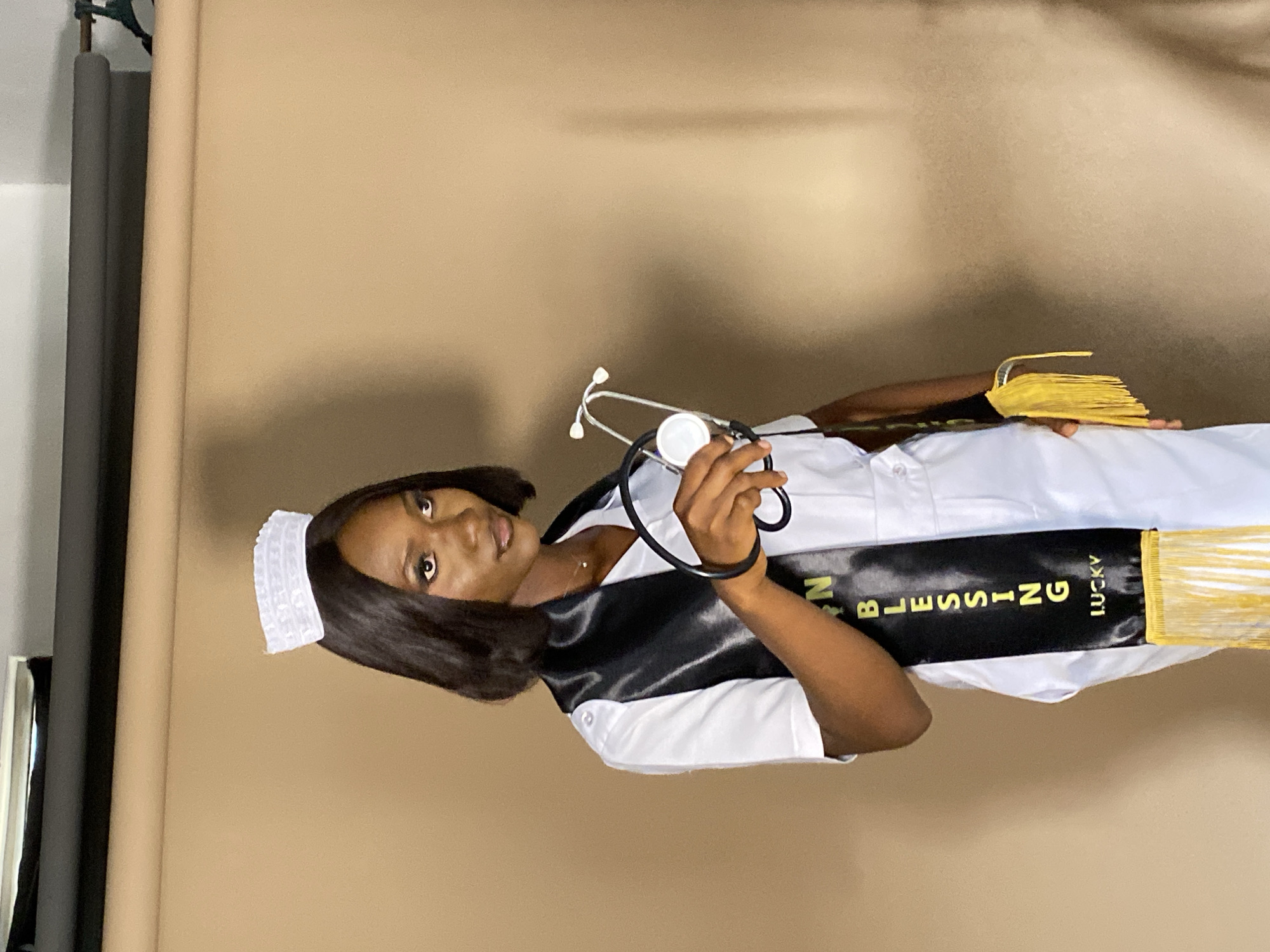 A woman dressed in a nurse's uniform is standing in front of a tan backdrop, likely in a studio setting. She is wearing a white nurse's cap and a black sash with yellow fringe that reads "RN BLESSING LUCKY." She is holding a stethoscope in her hands. The location appears to be Gura Top, Nigeria.
