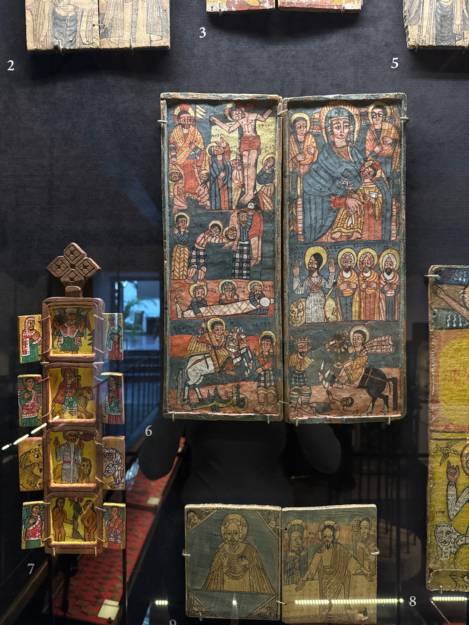 This image shows a close-up view of several religious icons displayed in a museum setting, likely an exhibition. The icons are small, rectangular wooden panels painted with intricate religious scenes and figures in a traditional style. They are arranged on a dark, textured wall, with white numbers indicating their order or cataloging.
The icons depict scenes from the Bible, including the Crucifixion of Jesus and the Virgin Mary with child. The style of painting is characteristic of Ethiopian art, with flat figures, bold outlines, and vibrant colors. Some icons are hinged, suggesting they might be part of a triptych or diptych.
The environment appears to be a museum with display cases or shelves. The lighting is subdued, with spotlights highlighting the icons. The overall impression is one of reverence and historical preservation.
The numbering visible in the image, particularly "2", "3", "5", "6", "7", "8", and "9", suggests a museum cataloging system. While the image itself does not provide direct clues to its specific location, if this is a part of a known exhibition or a photograph from a specific museum, that information would be necessary to pinpoint the city and country.
Without additional context or information beyond what is visually present in the image, it is impossible to definitively state that this image was taken in Moscow, Russia, or any other specific city. The scene is a generic museum display of religious art.