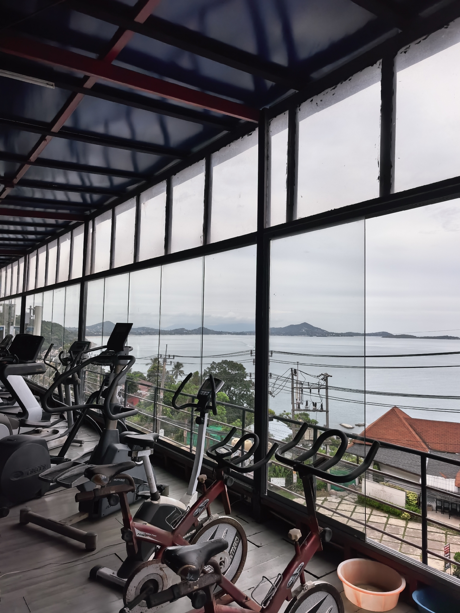 An indoor gym is depicted with a large glass window offering a view of the ocean and hills on a cloudy day. Several exercise machines, including treadmills and stationary bikes, are arranged in the foreground. The gym has a modern design with a metal and blue ceiling structure. Outside the window, power lines, trees, and buildings are visible, along with a red-roofed building. The scene appears to be located in a tropical or coastal area, possibly in Ko Samui District, Thailand, given the architecture and natural landscape.
