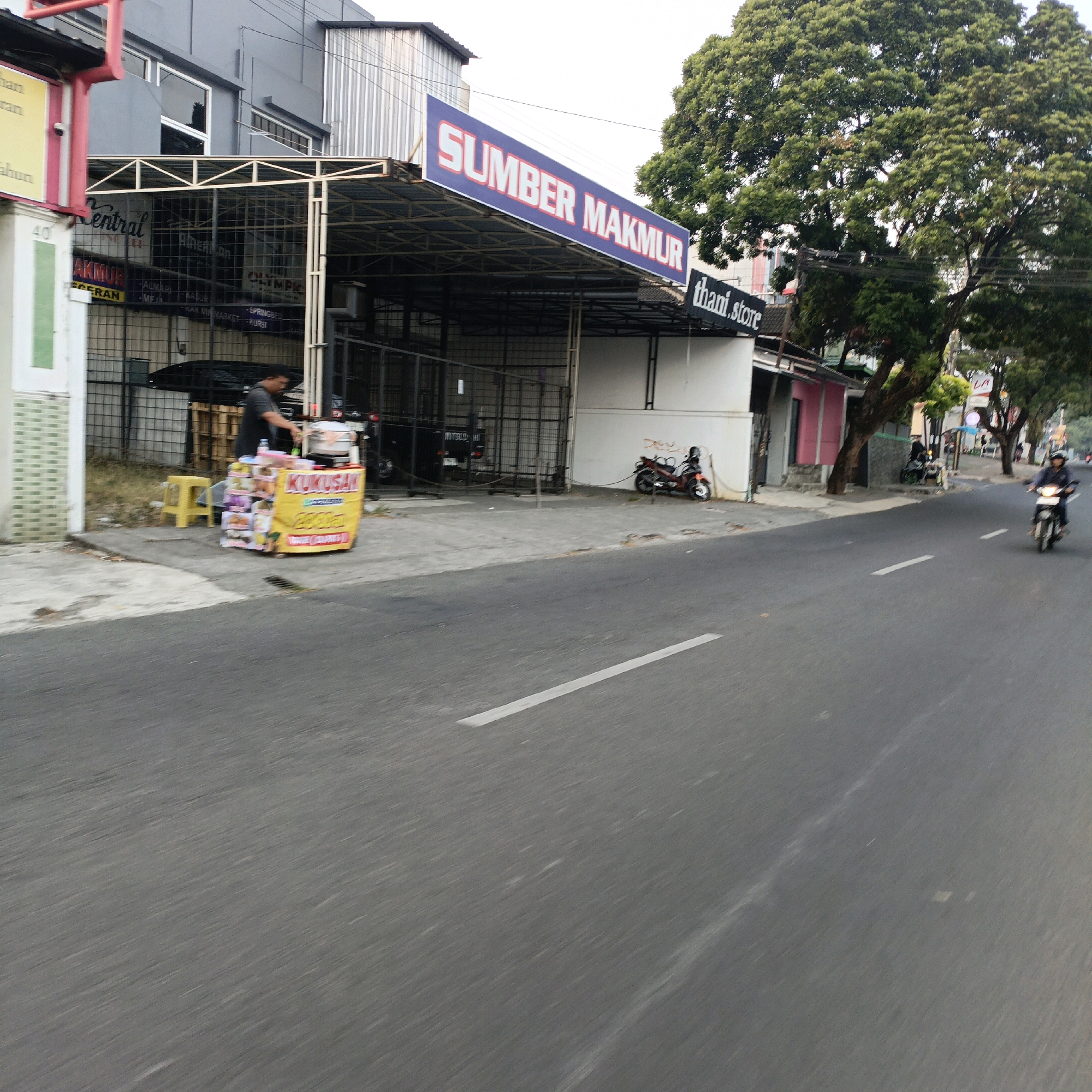 The image depicts a street scene in Salatiga, Indonesia. On the left, a building with a grey facade features a large blue and red banner with white lettering that reads "SUMBER MAKMUR". Adjacent to this is a black sign with white text stating "thani.store". A street vendor is operating a mobile food cart with a yellow banner displaying "KUKUSAN" and prices in Indonesian Rupiah. The cart is set up on the sidewalk. A man in a dark grey t-shirt is attending to the cart. A yellow plastic stool is placed beside the cart. Further down the street, a motorcycle is parked and tied to a pole. Several other motorcycles are visible, including one in motion driven by a person wearing a helmet, on the right side of the image. A large, leafy green tree dominates the right side of the frame. The road has a white line marking its center. The overall environment suggests an urban commercial area.
