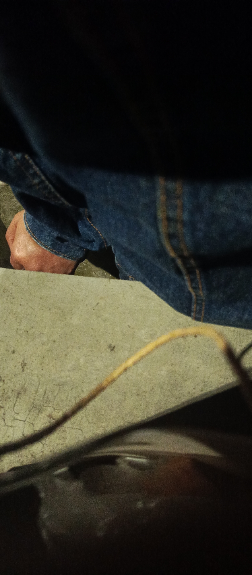 The image shows a close-up view of a person's hand, partially obscured by blue denim fabric. The hand is clenched into a fist and rests on a textured, light-colored surface, possibly concrete, which exhibits some fine cracks. The denim appears to be part of a jacket or shirt, with visible stitching. The lighting is dim, casting shadows and creating a somber atmosphere. The background is predominantly dark and out of focus. The specific location in Malang, Indonesia, cannot be determined from the visual information provided.