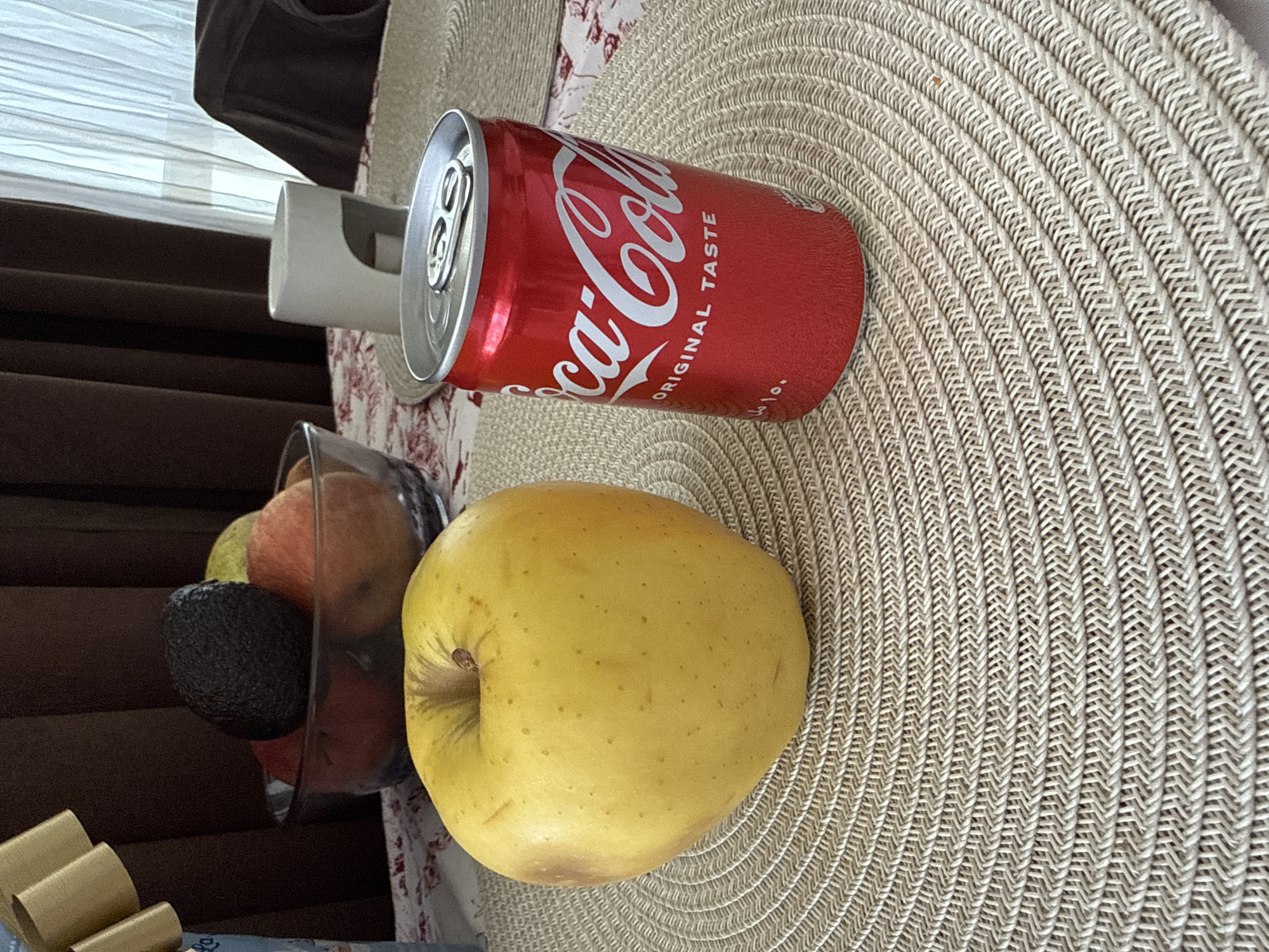 The image, taken in Blagoevgrad, Bulgaria, displays a still life arrangement on a tabletop, illuminated by bright daylight, likely from a nearby window.
The central subjects are a red Coca-Cola can and a large, smooth-skinned yellow apple, both resting on a light beige, circular woven placemat. The Coca-Cola can is prominently featured, displaying its iconic white "Coca-Cola" script and the words "ORIGINAL TASTE" on its red surface. Next to it, the yellow apple shows a few light brown blemishes and speckles.
To the left, a clear glass bowl holds an assortment of other fruits, including a dark, textured avocado and several reddish-orange fruits, possibly apples or oranges. The background features dark brown curtains and a window with white horizontal blinds, allowing natural light to enter the scene. A white, somewhat cylindrical object is partially visible behind the Coca-Cola can. A fabric with a red and white floral pattern can be seen beneath the placemat. In the bottom-left corner, a golden ribbon or bow from a package is partially visible, hinting at another item just outside the frame. The placemat itself exhibits a detailed, concentric woven pattern, adding texture to the surface.
No specific activity or event is taking place; the scene is a static arrangement of items. No people are visible.