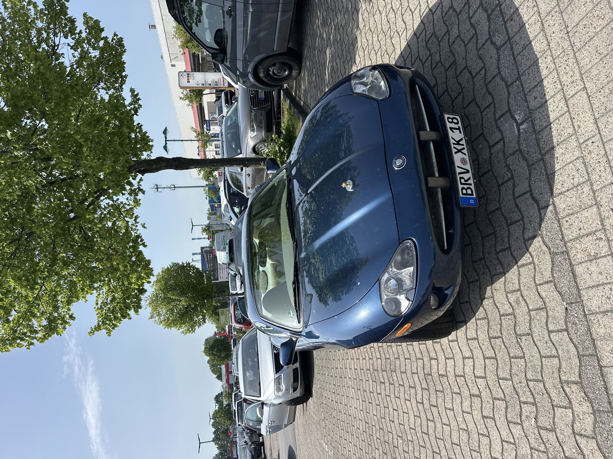 The image shows a parking lot in Ottersberg, Germany, with several parked cars. The closest car to the camera is a blue Jaguar XK with the license plate "BRV XK 18". The parking lot is paved with interlocking bricks. A tree partially obscures the view of the cars and buildings in the background. Other vehicles are visible in the row of parked cars. The sky is blue with some clouds.