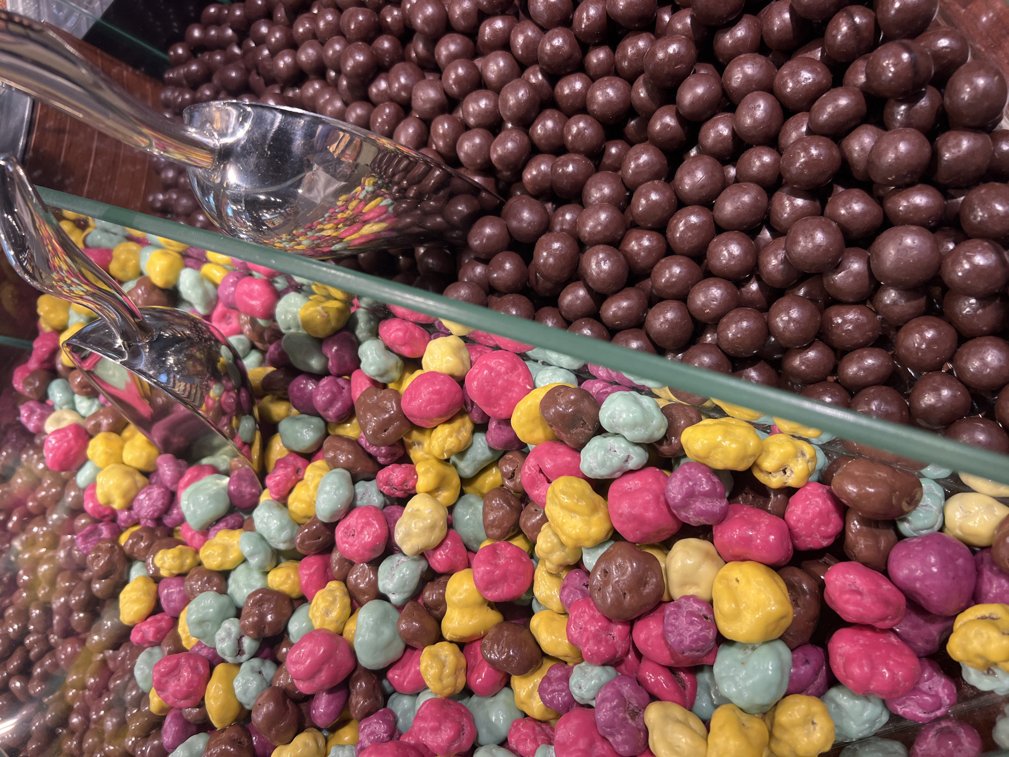 This media file is a close-up, eye-level shot of a display of chocolates. The foreground is filled with brightly colored, irregularly shaped candies, including pink, yellow, light blue, and dark brown varieties. These candies are likely covered in a hard candy shell and filled with chocolate. In the background, there is a large pile of smaller, uniformly round, dark brown chocolates, which appear to be chocolate-covered raisins or nuts.
Two silver scoops are visible in the display. One scoop is in the upper left corner, partially submerged in the dark brown chocolates, and reflects the colorful candies below. The second, smaller scoop is in the lower left, resting on the colorful candies. The candies are arranged in clear plastic or glass containers, suggesting a retail or confectionery setting, possibly a candy store or a section of a grocery store. A green-colored shelf or divider separates the two types of chocolates.
The lighting is bright, suggesting an indoor setting, possibly during the daytime. There is no visible text in the image. The overall impression is one of abundance and variety, with a focus on sweet treats. The location context provided is Odesa, Ukraine, which, combined with the visual cues, suggests this is likely a confectionery shop or market stall in Odesa. The image doesn't convey any specific time of day or weather conditions beyond the indoor lighting.