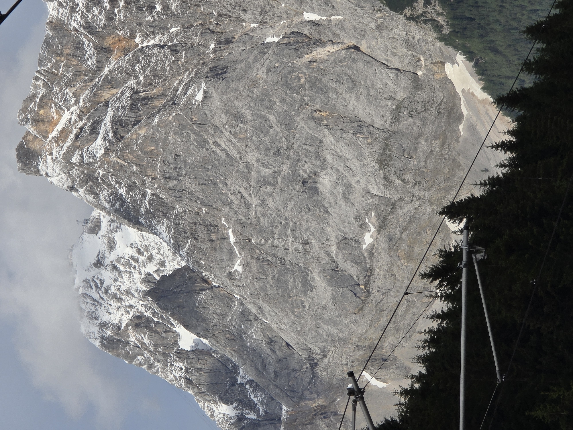 The image captures a majestic mountain landscape in Lermoos, Austria. The towering peaks are partly covered in snow, creating a stark contrast against the grey rock face. The foreground is dominated by dark green pine trees and the infrastructure of a cable car system, with its wires and supporting structures visible. The cable car wires stretch across the frame, leading towards the imposing mountains. The sky is a mix of blue and grey clouds, hinting at varied weather conditions. The scene evokes a sense of natural grandeur and the presence of human engineering in a wild environment.