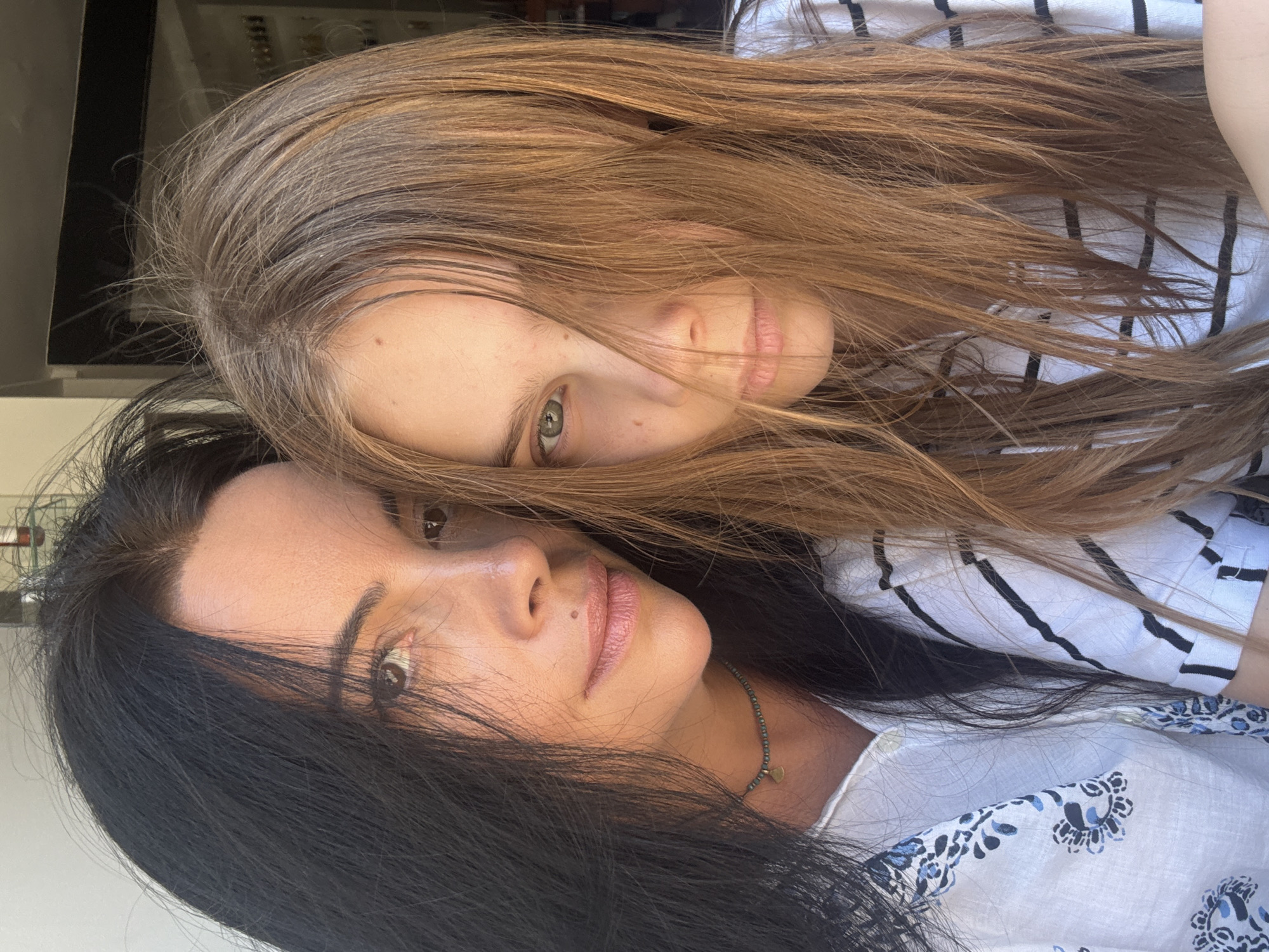 A close-up selfie taken outdoors shows two females, one adult and one adolescent, positioned in the foreground. The adult, on the left, has long black hair and is wearing a white blouse with a blue floral pattern and a beaded necklace. The adolescent, on the right, has long auburn hair, which partially obscures her face, and is wearing a white shirt with black stripes. The background is blurred, with hints of a sunlit urban environment, possibly a balcony or outdoor seating area. The lighting suggests it is daytime. No specific landmarks identifying Tel-Aviv are visible.