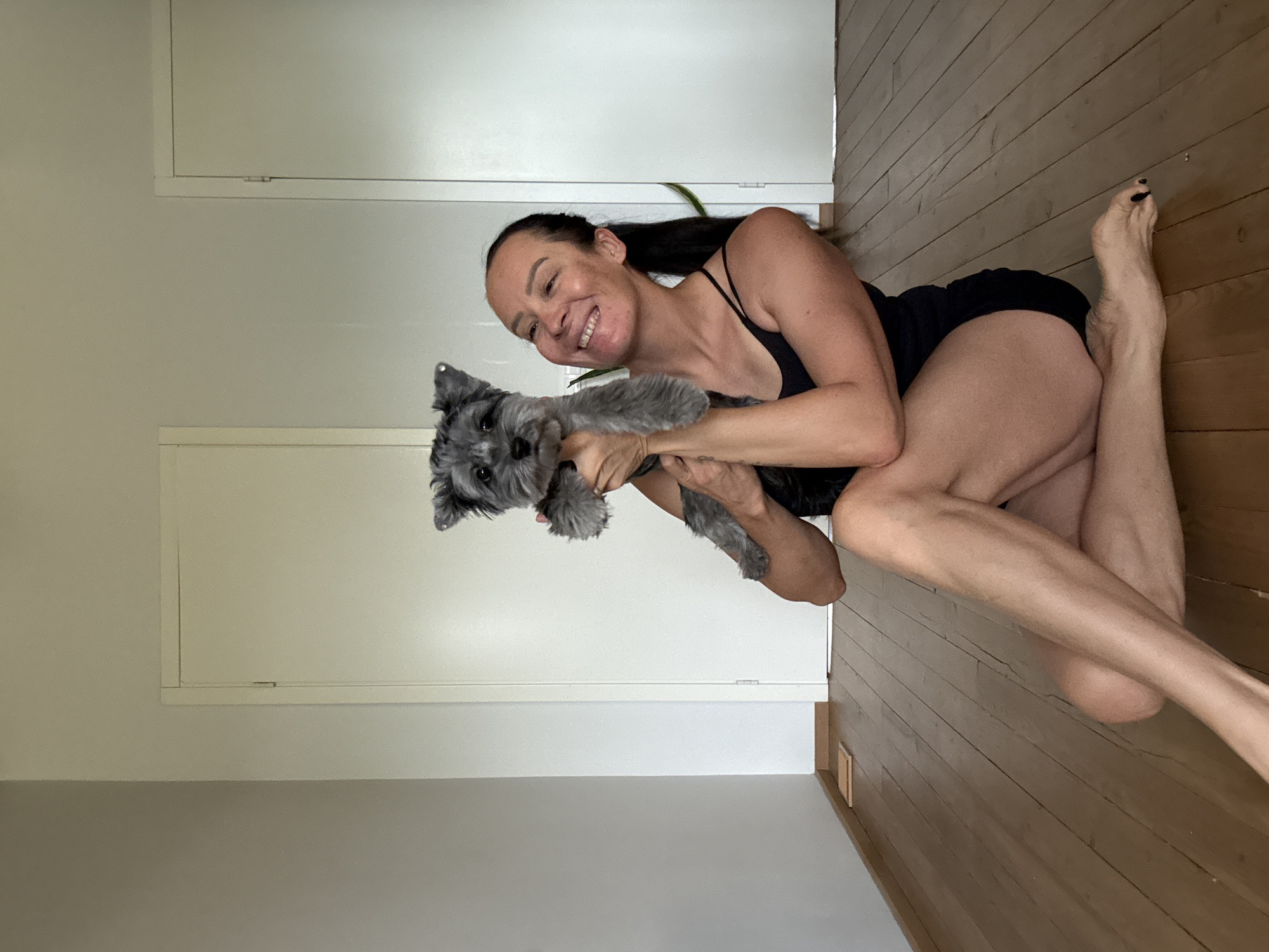 A woman with dark hair pulled back in a ponytail and a black tank top is sitting on a wooden floor. She is holding a small, gray dog in her arms. The dog is facing the camera. The woman is smiling. She is wearing black shorts. Her legs are crossed. The floor is made of wood planks. In the background, there are two white doors and a light-colored wall. The scene appears to be indoors. The image does not provide any information to suggest the location is Chaikovsky, Russia.