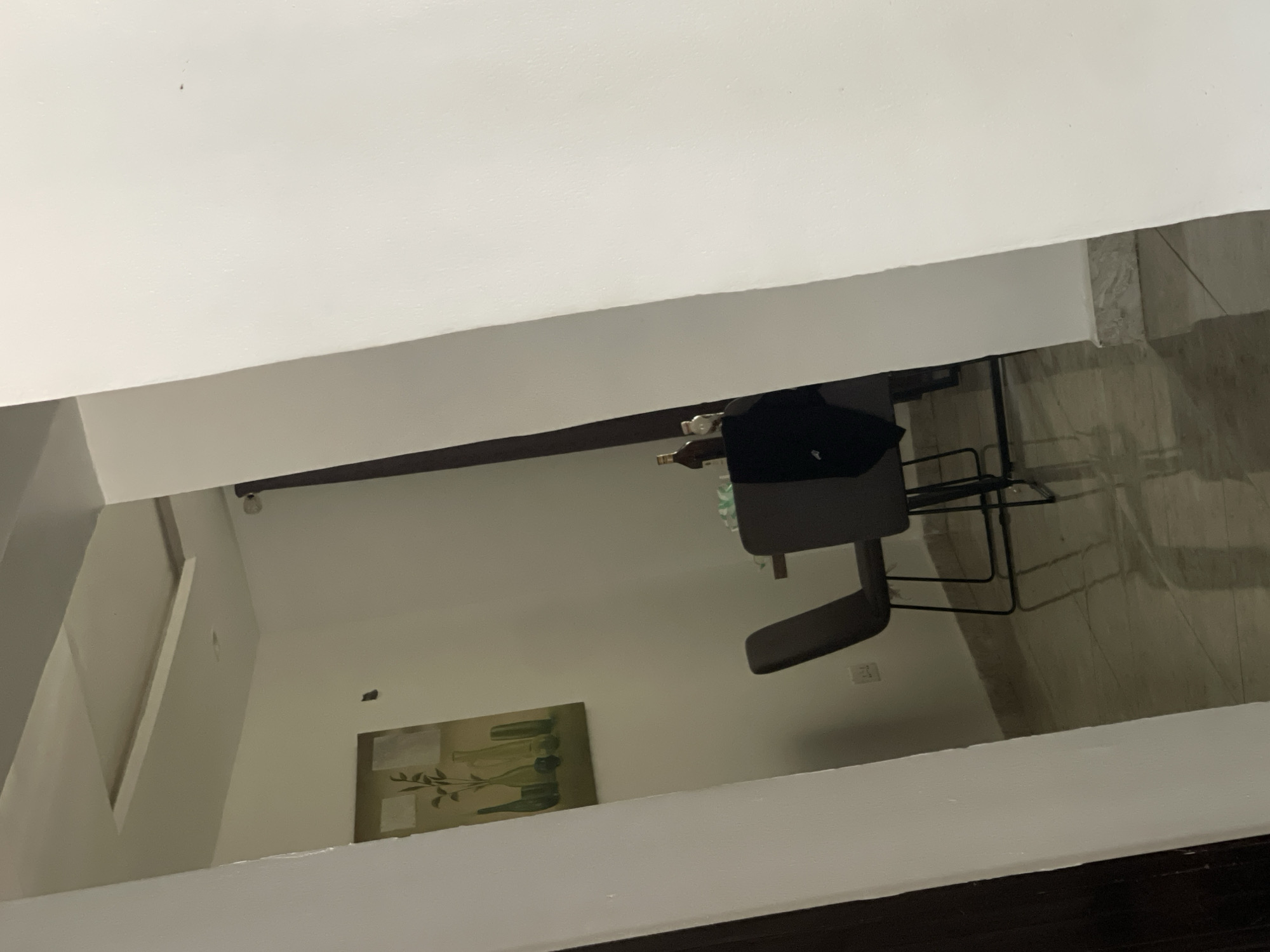 The image appears to be taken from a high vantage point, possibly through a doorway or window. The view looks into a room with white walls and tiled floors. A gray office chair is visible near a dark table or desk. A picture hangs on the wall. The room seems to be in a residential or office space. The location in Iddo Tudunwada, Nigeria cannot be confirmed from this image. No people are visible.