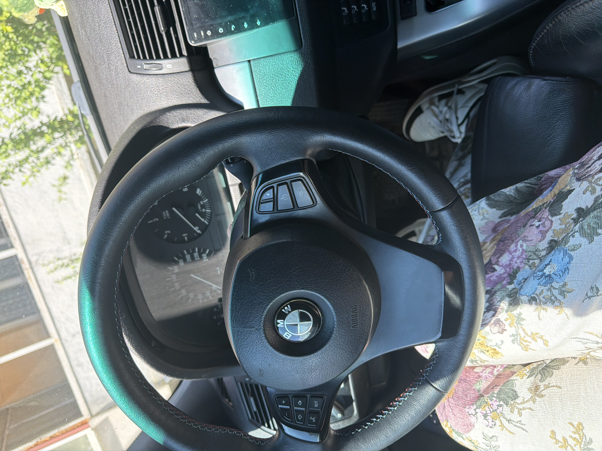 This media file showcases the interior of a car, specifically focusing on the steering wheel and dashboard area. The steering wheel is black leather with red and blue stitching around its rim. In the center of the steering wheel is the BMW logo. Various buttons are visible on the steering wheel, including those for cruise control and other functions. The dashboard includes an instrument cluster with a speedometer and tachometer, and an air vent. A portion of a digital screen is also visible above the dashboard.
In the background, a pair of white athletic shoes are visible, partially obscured by a floral-patterned fabric. Sunlight streams into the car, casting shadows and highlighting the textures of the interior. The overall impression is that of a parked car on a sunny day, likely in an outdoor setting given the natural light and the hint of greenery visible through the windshield. No specific activity or event is depicted, but the scene suggests a moment of pause or readiness for a journey.
The image is oriented such that the top of the frame appears to be on the left, suggesting it was taken with the camera held sideways. No discernible text is present in the image other than the branding on the steering wheel and the "AIRBAG" label. The context of Pazardzhik, Bulgaria, provides a geographical location, but no specific visual cues within the image directly confirm this. The lighting suggests it is daytime.