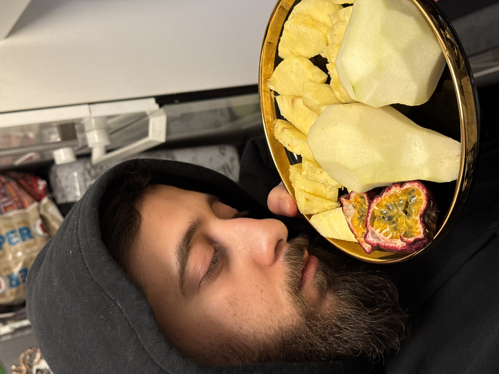 The image depicts a close-up, vertical shot of an adult male in an indoor setting within Strasbourg, France. The male, positioned on the left side of the frame, has dark hair, a prominent beard, and is wearing a black hooded sweatshirt with the hood pulled over his head. His eyes are closed or cast downwards intently, focusing on a black plate with a gold rim held in his right hand. The plate contains various cut fruits: numerous yellow, triangular pieces of pineapple, two halves of a red-skinned passion fruit displaying its yellow pulp and dark seeds, and two large, peeled, pale-green fruit halves, possibly pears or apples. The male's hand is visible holding the plate from underneath. The background is out of focus, revealing a white household appliance on the right, possibly a refrigerator or cabinet, and blurred items, including what appears to be a beige bag with red and blue lettering, behind and above the male's left shoulder.