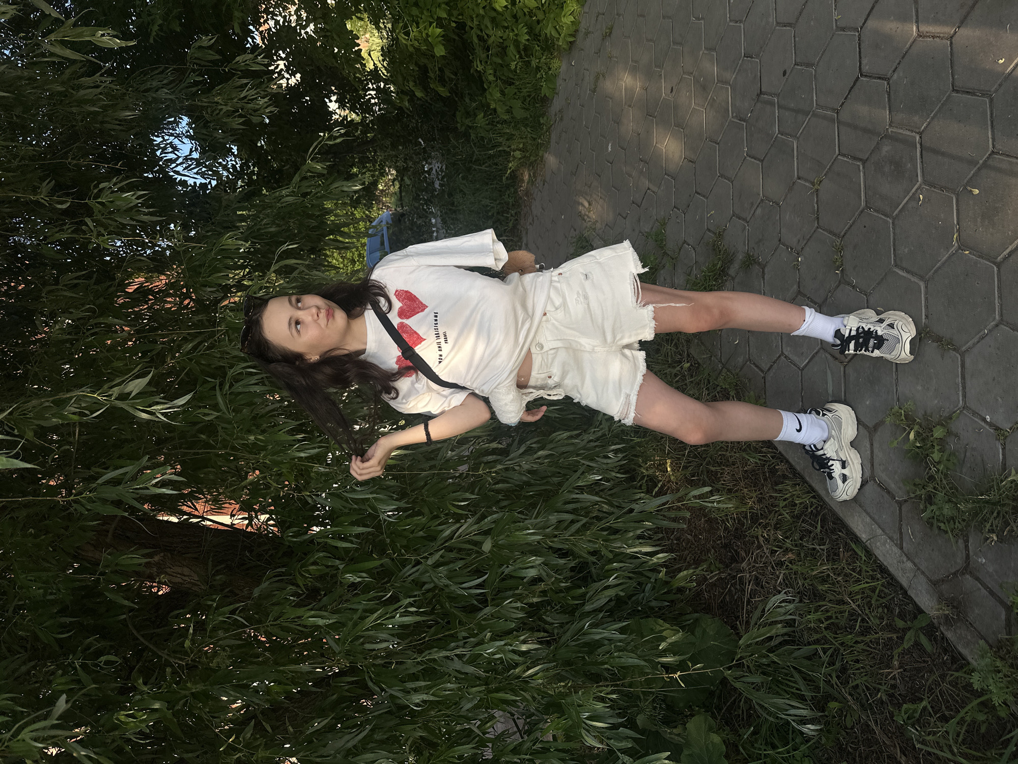 A young woman with dark hair is standing on a paved walkway under a large tree. She is wearing a white t-shirt with red hearts on it, white distressed denim shorts, white socks with a black Nike logo, and chunky sneakers. She is holding a strand of her hair and looking to her right. She has a small furry bag slung over her shoulder. The walkway is made of hexagonal paving stones. There is green foliage from the tree and other plants surrounding the walkway. In the background, there is a hint of buildings and a car. The lighting suggests it is daytime, likely late afternoon given the shadows. This scene is located on a street in Votkinsk, Russia.