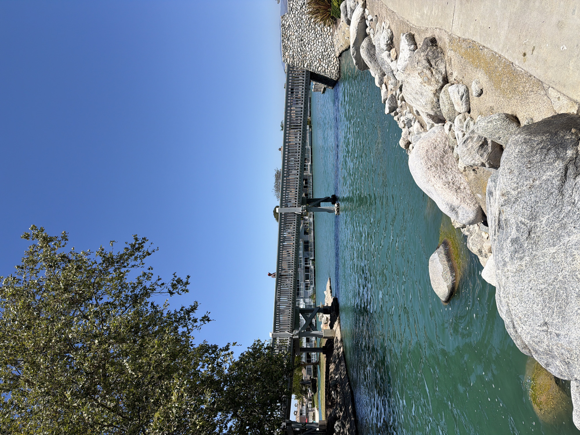 The image depicts a canal with turquoise water in Irvine, California, United States of America. A wooden pedestrian bridge spans the canal, with a single person standing on it. The bridge is supported by concrete pillars. On the right bank of the canal, there is a pathway lined with rocks and sand, and a wall constructed of large stones. Lush green trees are visible on the left bank, casting shadows on the ground. In the background, buildings with white facades and red-tiled roofs suggest a residential or resort area. The sky is a clear, bright blue.