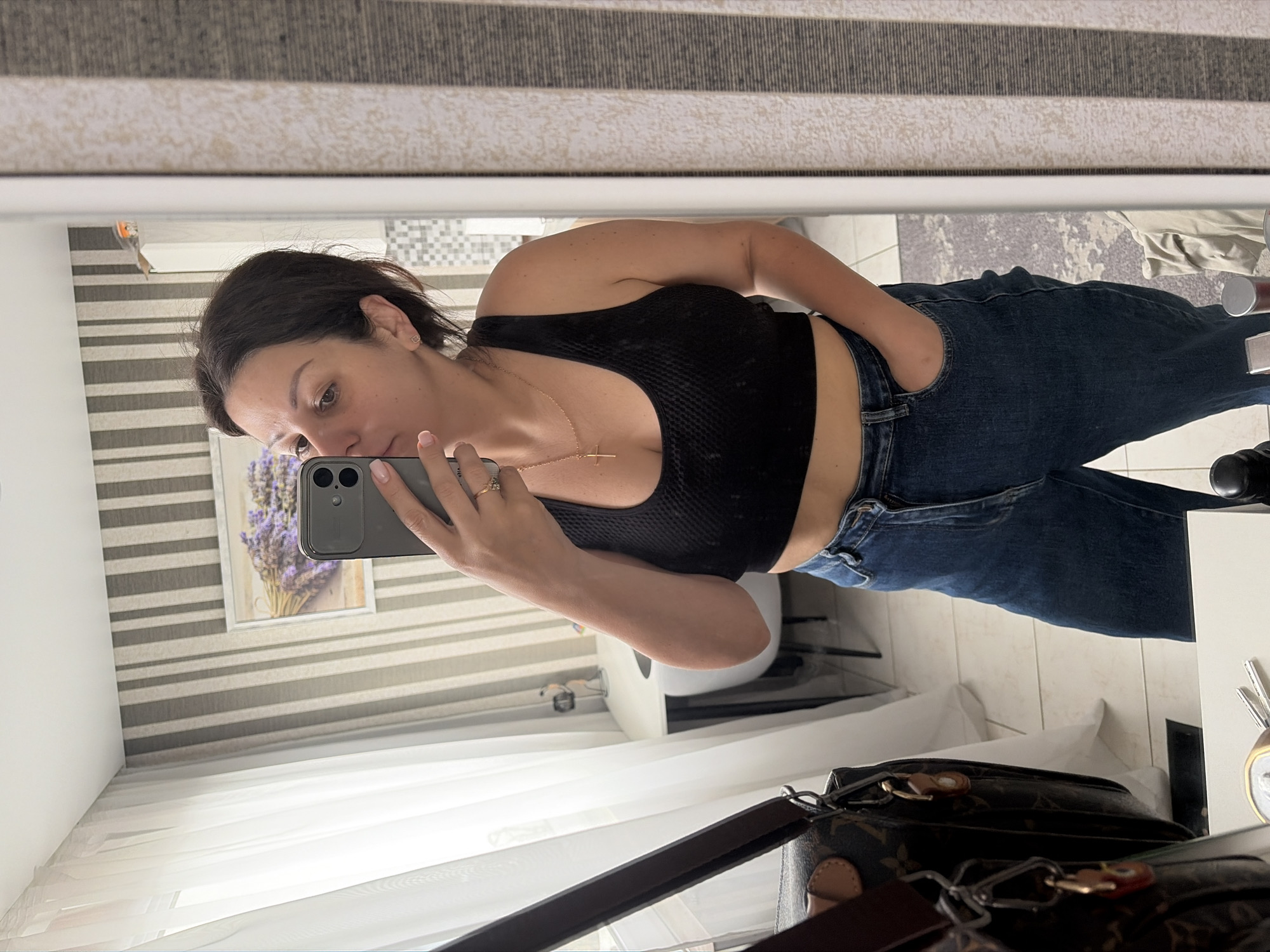 The media file shows a young woman taking a mirror selfie in a room. She is wearing a black crop top and blue jeans. She has dark hair and is looking at her phone, which she is holding in her left hand. Her right hand is in her pocket. There is a cross necklace around her neck. In the reflection of the mirror, we can see a framed picture of lavender flowers on the wall, striped wallpaper, and a window with white curtains. A designer handbag is visible in the foreground. The overall impression is of a casual moment captured at home. No text is visible in the media file.