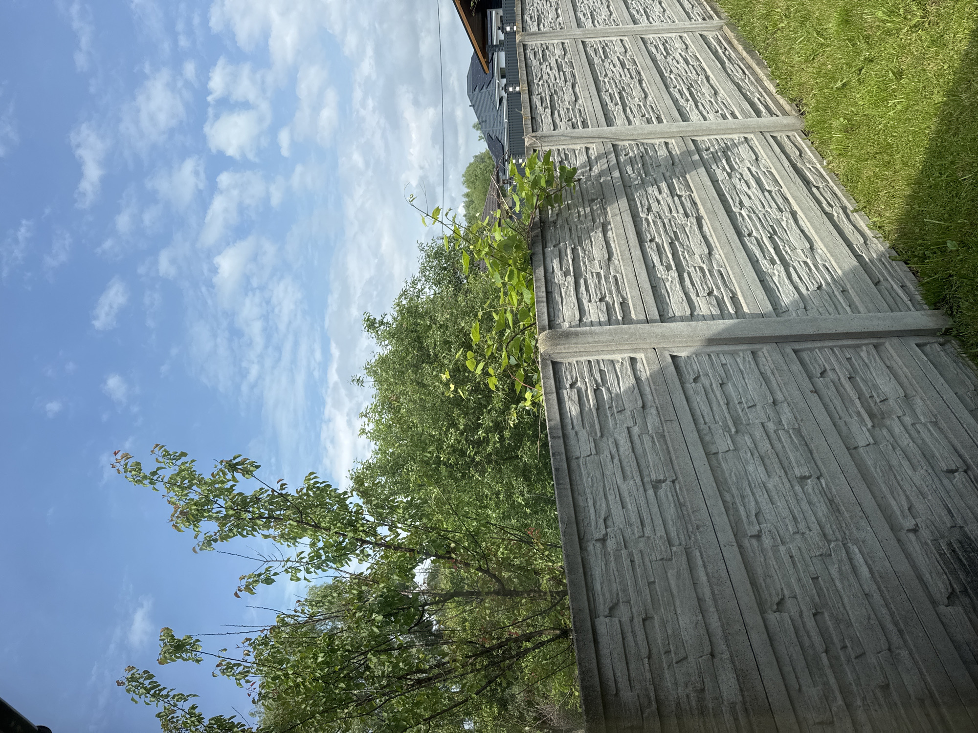 The image displays an outdoor scene, likely a residential area, under a bright blue sky with scattered white clouds. The primary subject is a grey pre-cast concrete fence running diagonally from the lower left to the middle right, dividing the foreground from the midground. The fence panels feature a textured, horizontal stacked-stone or slat design, separated by smooth vertical concrete posts. Green vegetation, specifically a leafy vine, grows over the top of the fence on the right side.
In the immediate foreground, to the right of the fence, is a patch of bright green grass, partially illuminated by direct sunlight and partially shaded.
Behind the fence, a dense array of green trees and shrubs occupies the left and central midground. Their foliage extends above the fence line and into the sky. In the midground-to-background on the right, several house roofs are visible. One prominent roof is dark grey or black, fitted with multiple solar panels. Adjacent to it, a portion of a brownish-orange roof with visible eaves and a gable is seen. Thin, dark utility lines are discernible against the sky above these structures.
The lighting indicates a sunny day, with shadows cast towards the right, particularly on the fence and grass. No individuals or animals are present in the frame. The location is in the city of Unknown, Unknown.