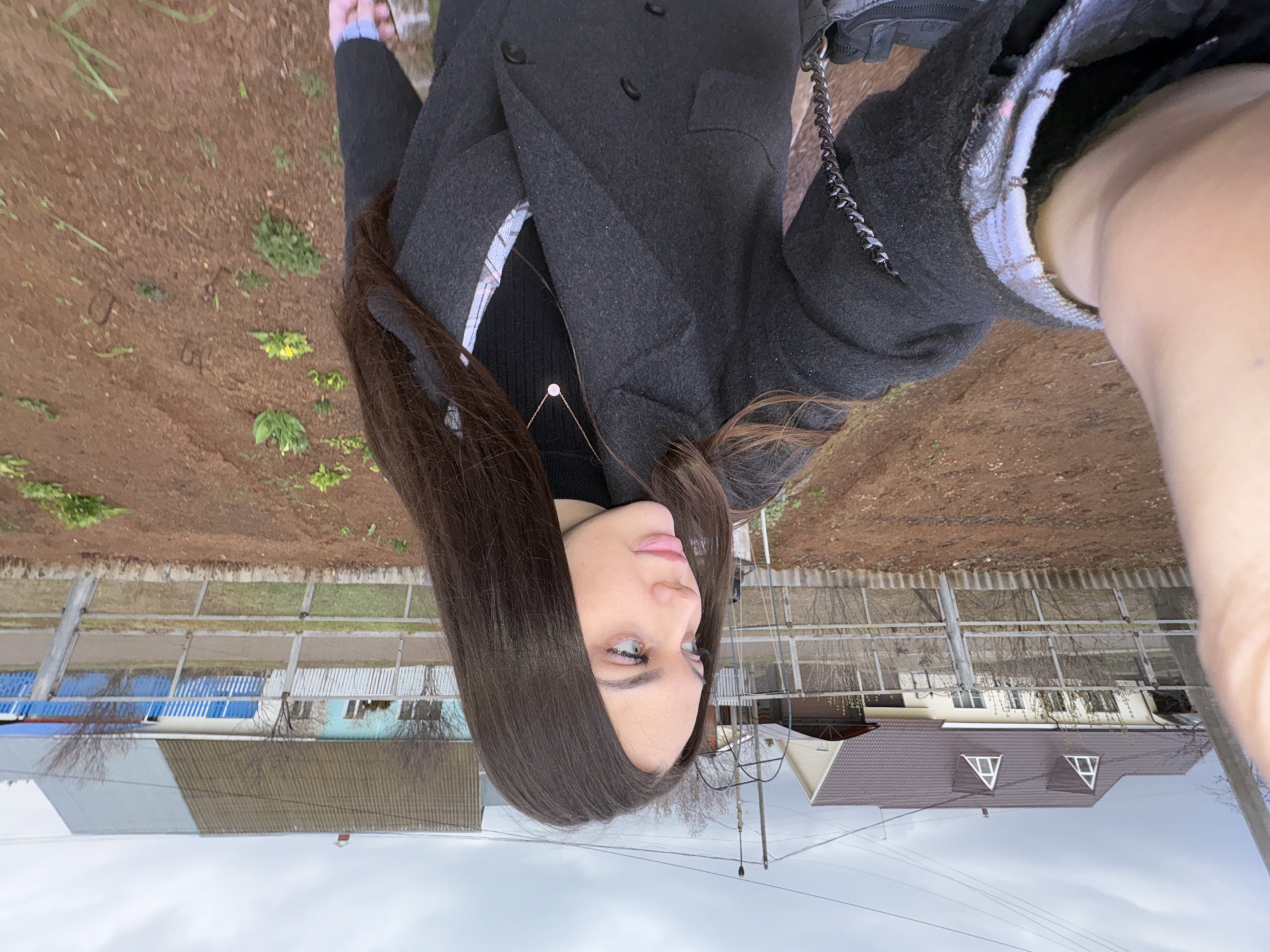 A young woman with long, straight, dark brown hair and light-colored eyes is featured in a self-portrait, oriented with her head on the left looking towards the right. She wears a dark grey double-breasted coat over a black top, with a glimpse of a plaid shirt underneath and a delicate gold necklace with a small pendant. A black bag with a chain strap is visible on her left side.
The setting is outdoors, likely a residential or semi-rural area in Прикамский, Russia. The immediate foreground consists of brown earth with sparse green patches and budding plants, indicative of early spring. In the midground, a fence line separates the viewer from several buildings, including a house with a blue roof and light blue walls, and another with a dark brown pitched roof featuring two triangular dormer windows. Bare trees and utility poles with power lines are visible against an overcast, light grey sky.
The woman is posing for the camera with a calm, neutral expression. The diffused light suggests a cloudy day. No visible text is present in the image.